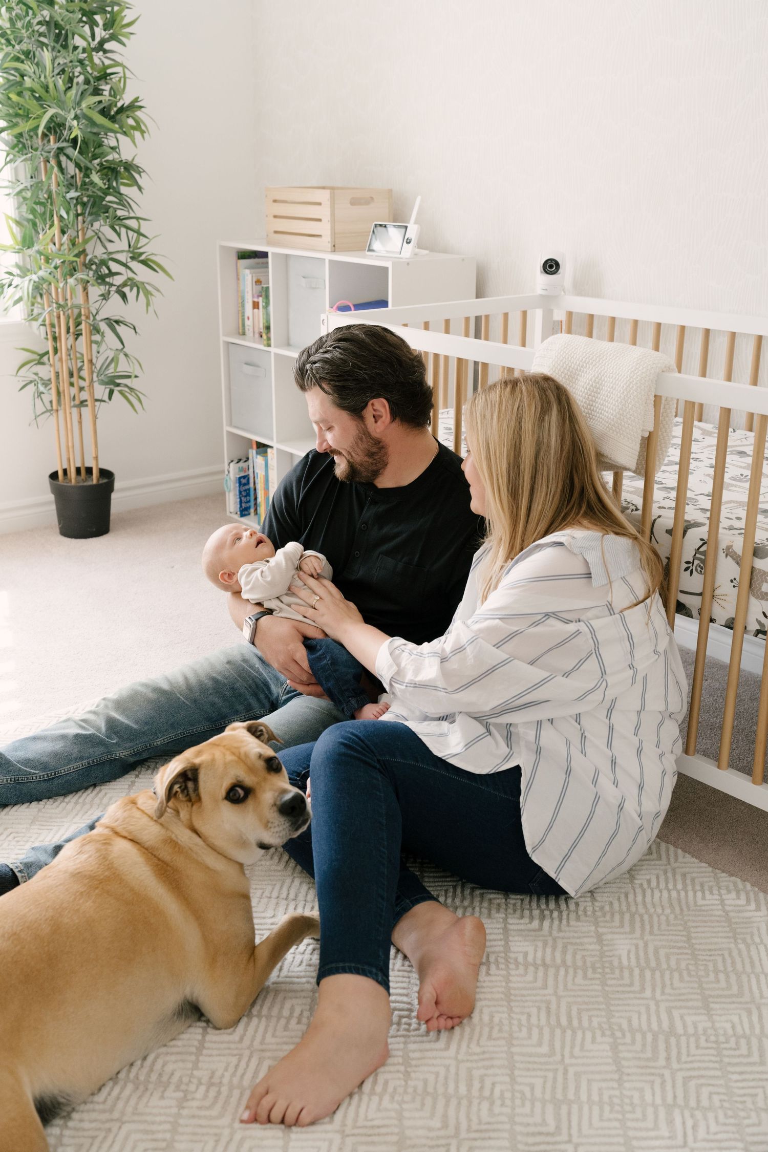Young family with newborn baby and golden dog sitting on nursery floor near white crib.