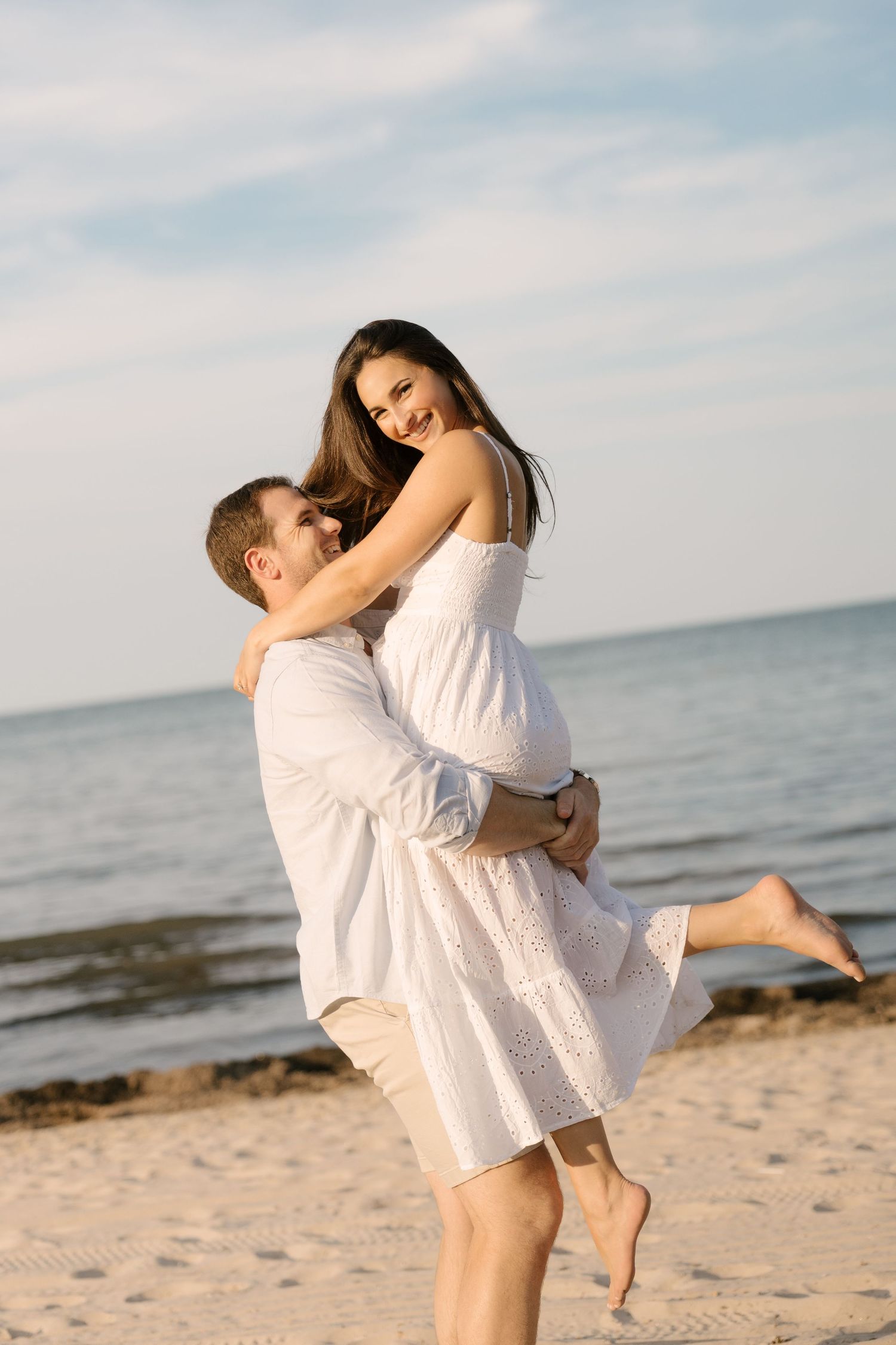 Happy couple on beach, man lifting laughing woman in white eyelet dress with ocean and blue sky behind them.