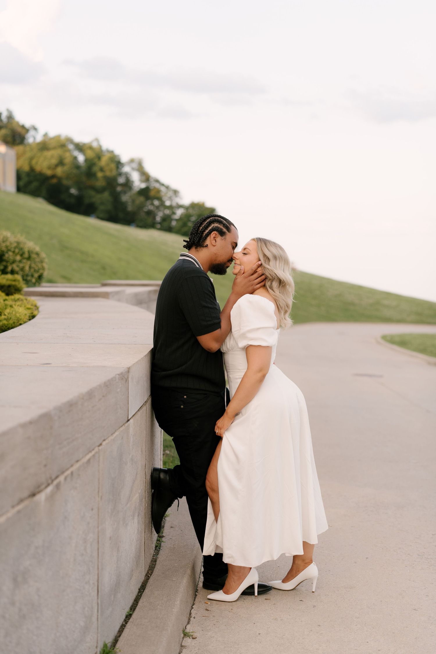 Couple sharing romantic kiss against concrete wall outdoors, woman in white dress with slit and heels.