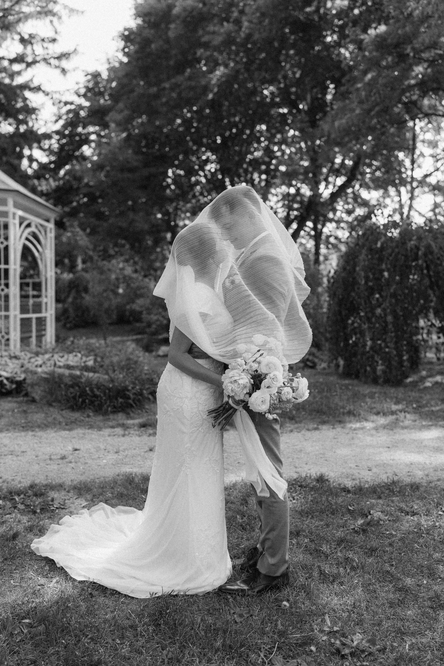 Black and white photo of a bride and groom kissing under a billowing veil in a garden setting.