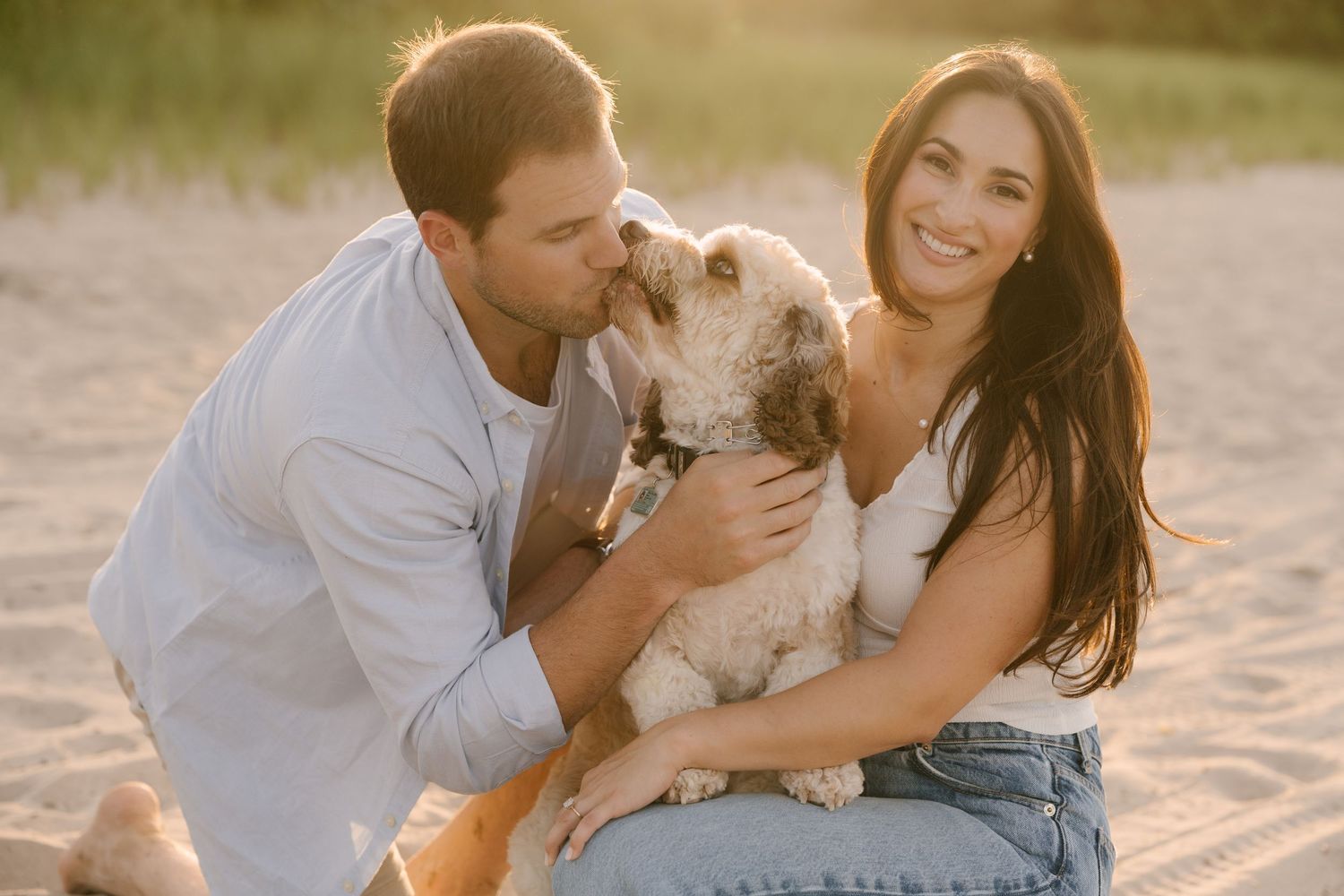 Smiling couple sitting on a sandy beach at golden hour with their fluffy white dog.