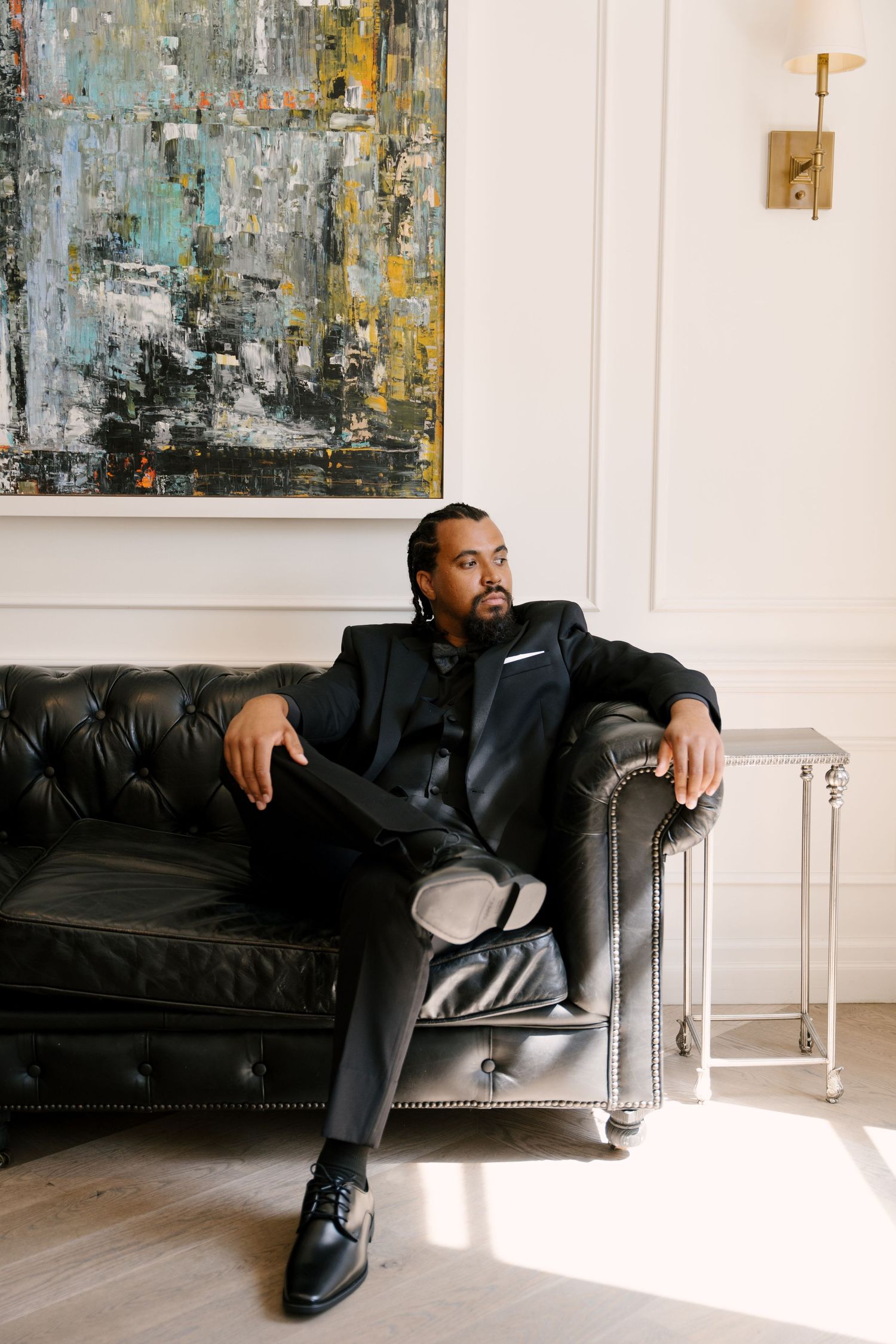 Dapper man in black suit sitting on leather sofa beside abstract wall art in The Walper Hotel.