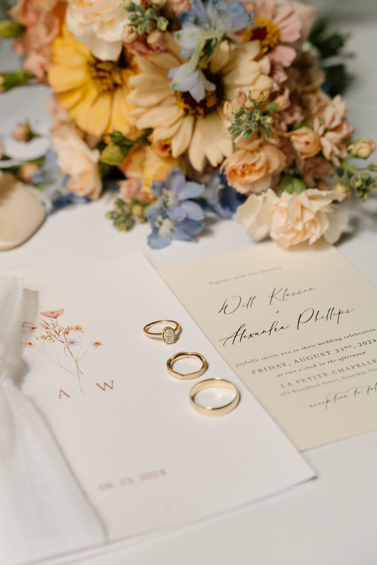 Gold wedding rings rest on elegant wedding invitations beside a colorful bridal bouquet of yellow, peach, and blue flowers.
