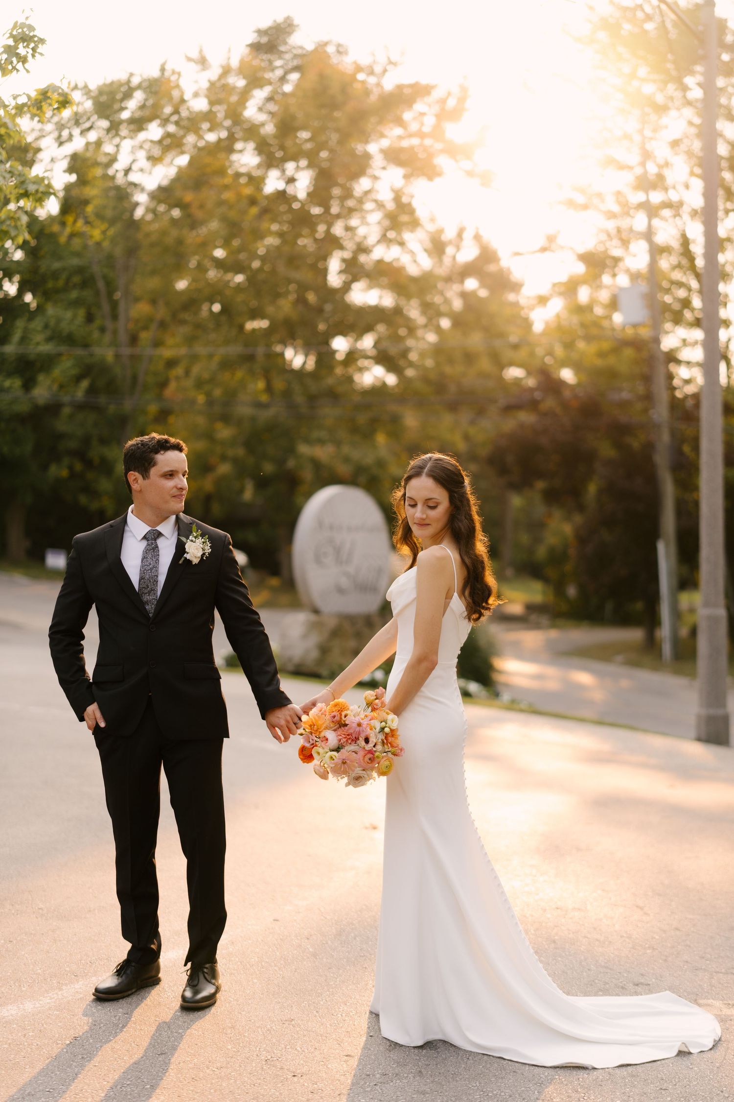 Bride and groom hold hands at golden hour, she in a white gown with bouquet, he in a black suit in front of Ancaster Mill venue sign.