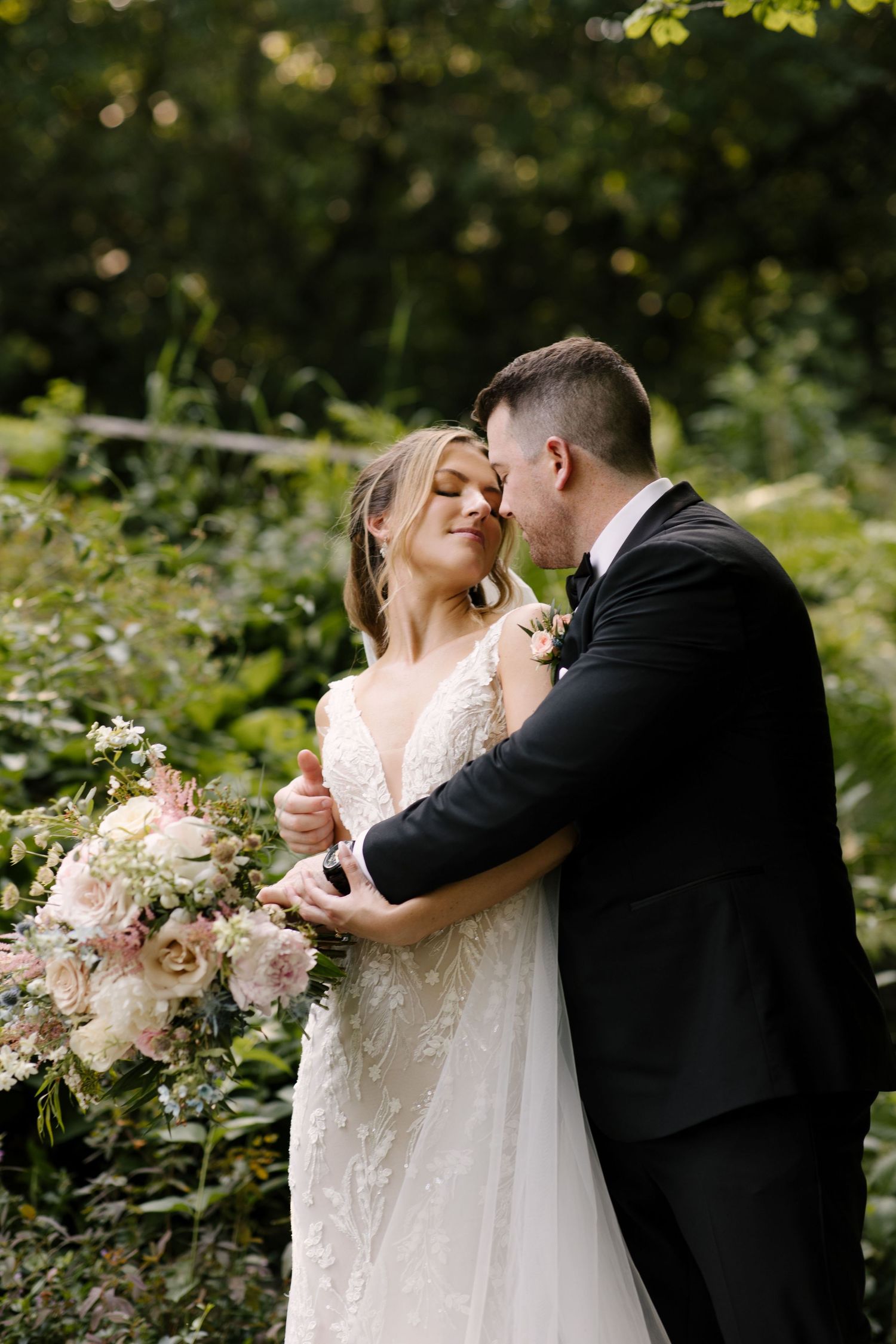 Bride and groom embracing in a lush green garden, bride holding a large blush floral bouquet.