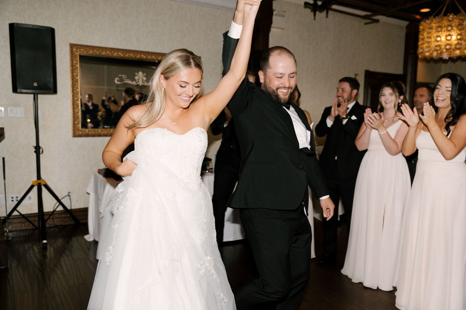 Bride and groom celebrate on the dance floor, hands raised, surrounded by cheering guests at their wedding reception.