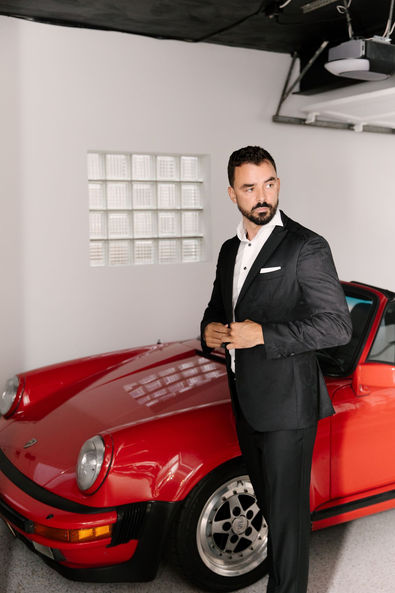 Groom in black tuxedo poses beside a classic red Porsche sports car in a white garage.