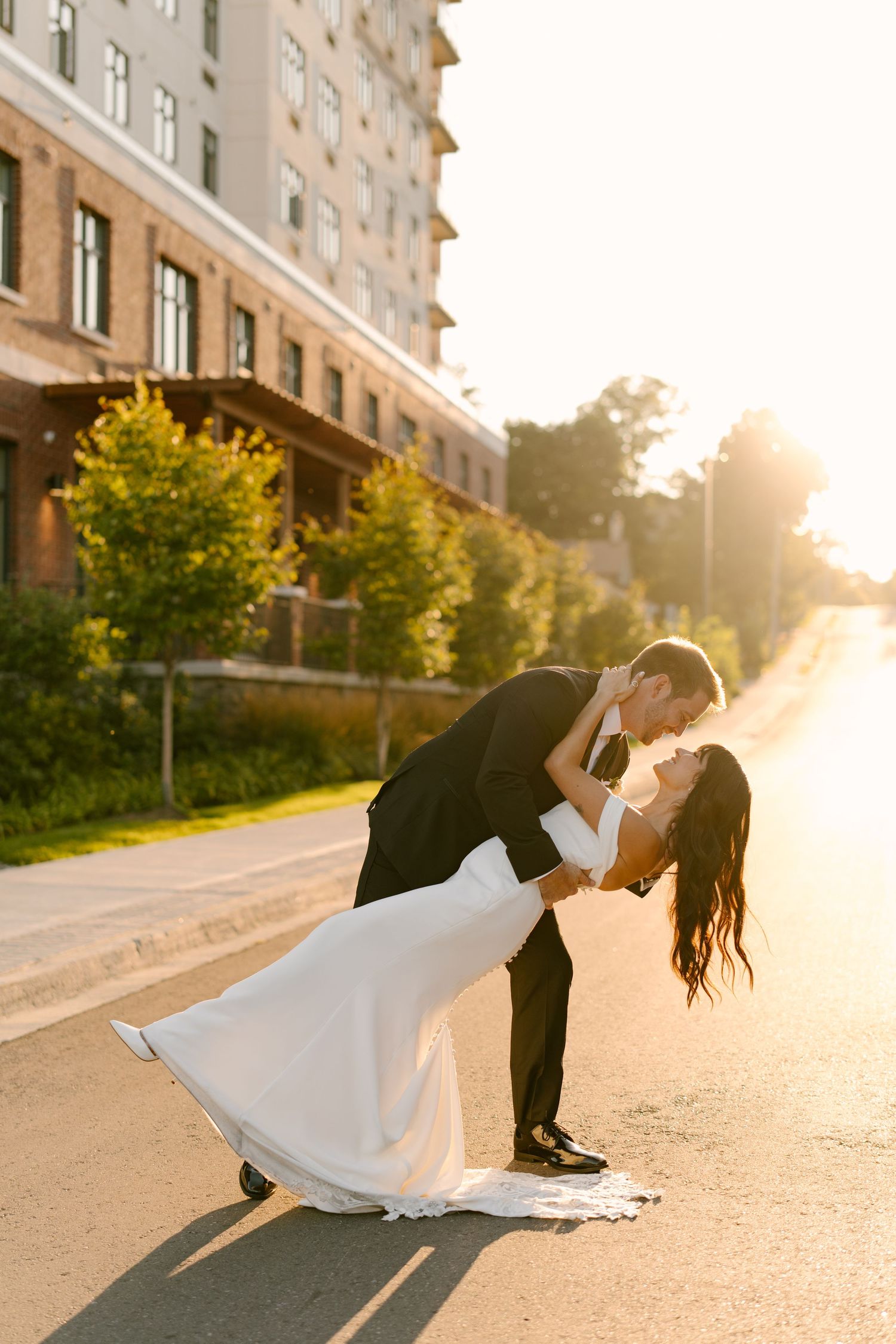 Groom dipping bride in romantic kiss on sunlit street, her white gown flowing in beautiful golden hour light.
