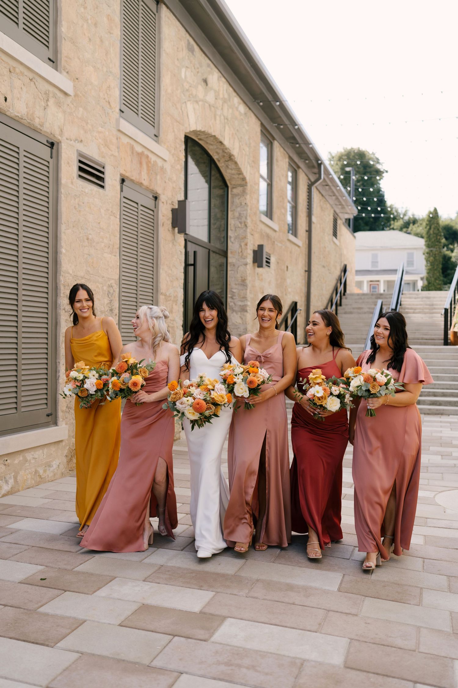 Bride in white gown walks with five bridesmaids in mismatched warm-toned satin dresses holding colorful bouquets.