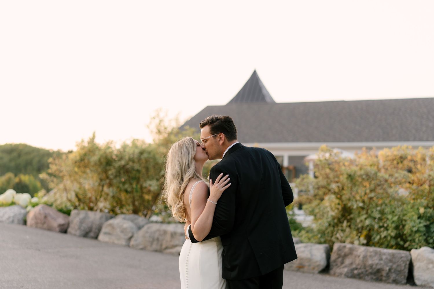 A bride and groom share a romantic kiss outdoors at golden hour with a elegant Whistle Bear wedding venue building in the background.