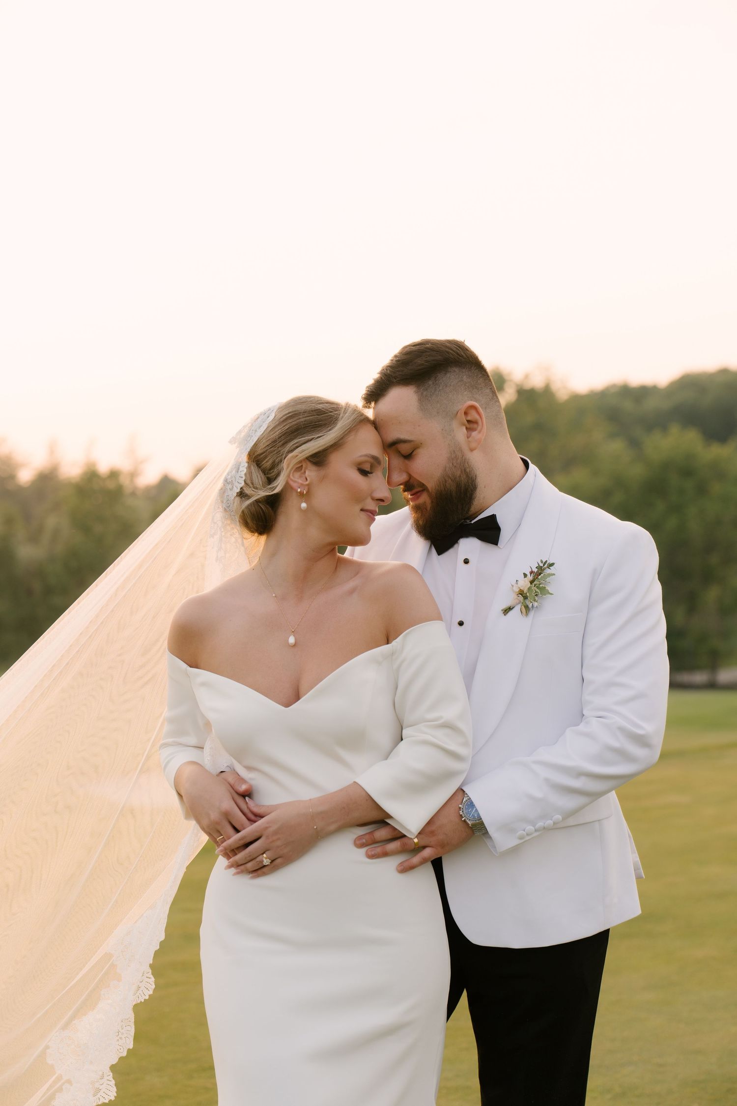 Romantic bride and groom embracing tenderly in golden sunset light, groom in white tuxedo jacket with bow tie outside Whistle Bear.