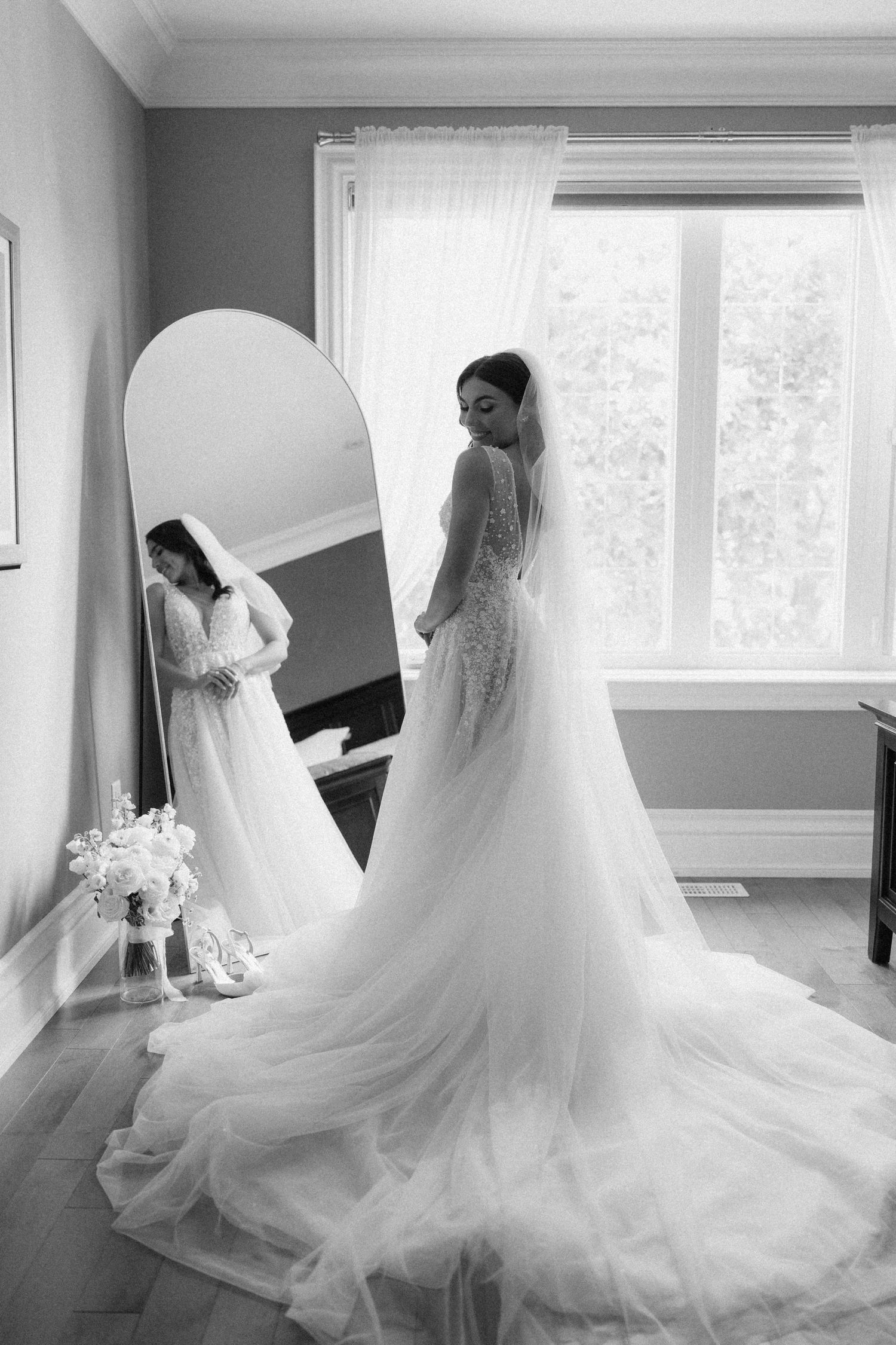 Black and white photo of bride in full lace gown with cathedral train standing before an arched mirror.
