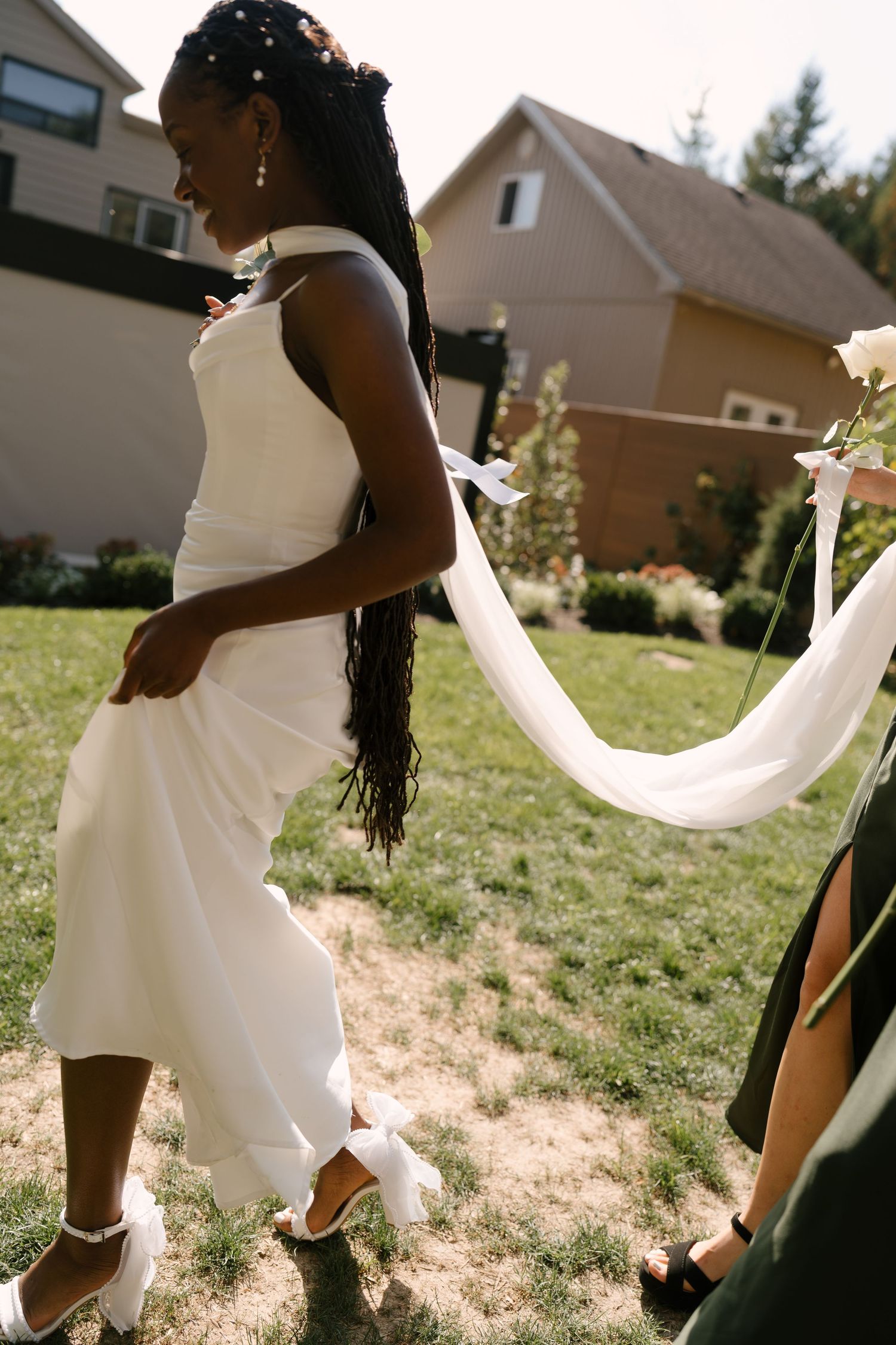 Black bride in white midi dress walking outdoors with bridesmaid holding her flowing veil train.