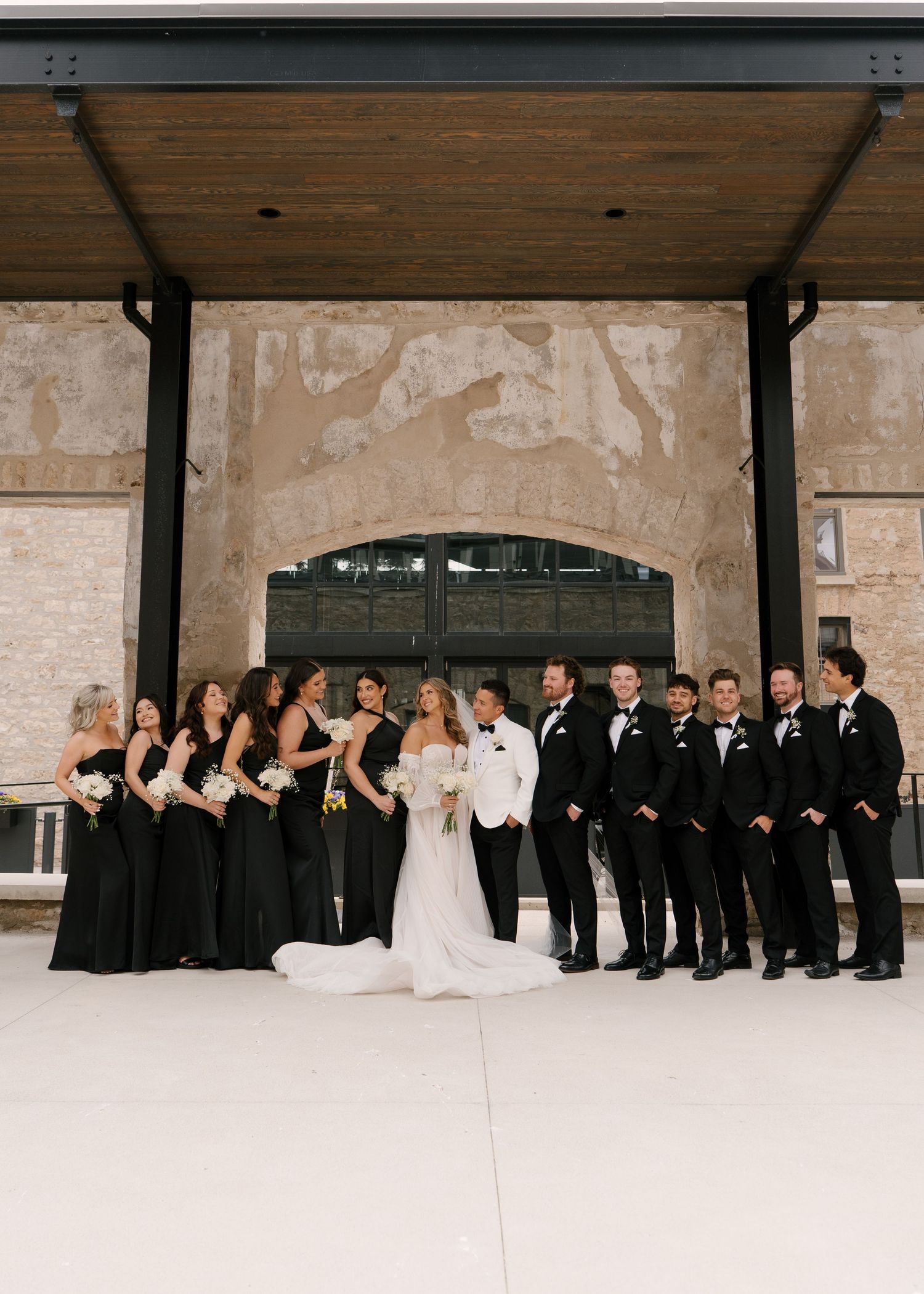 Bride and groom with large wedding party in black dresses and suits at stone Tapestry Hall wedding venue.