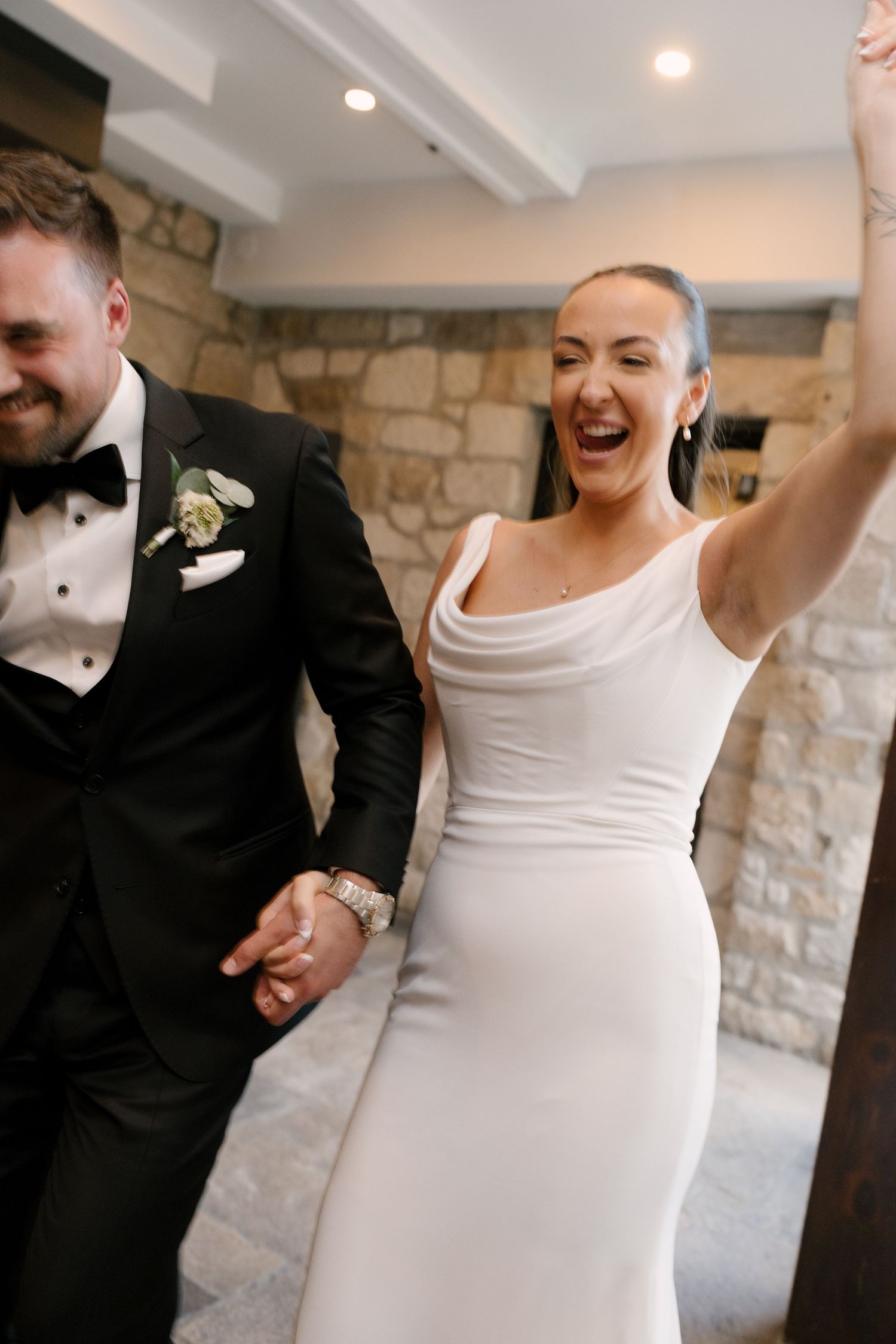 Joyful bride in white cowl-neck dress raising arm while entering reception with groom in black tuxedo.