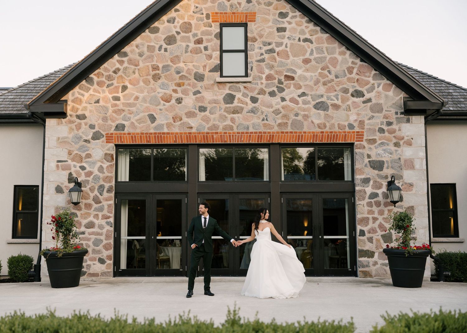 Bride and groom holding hands in front of a rustic stone and white wedding venue building at dusk at Roseville Estates wedding venue.