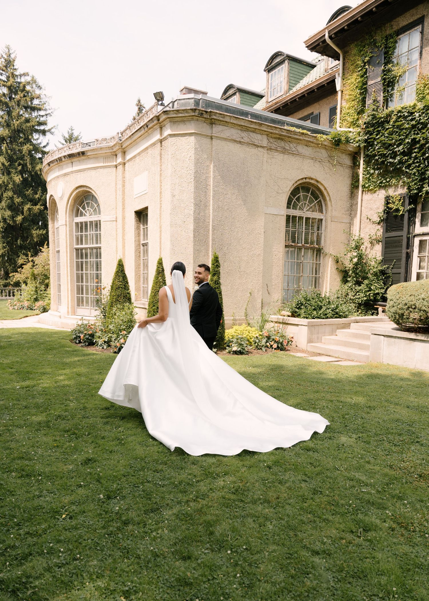 Bride in flowing white gown with long train stands with groom outside elegant ivy-covered Parkwood estate venue.