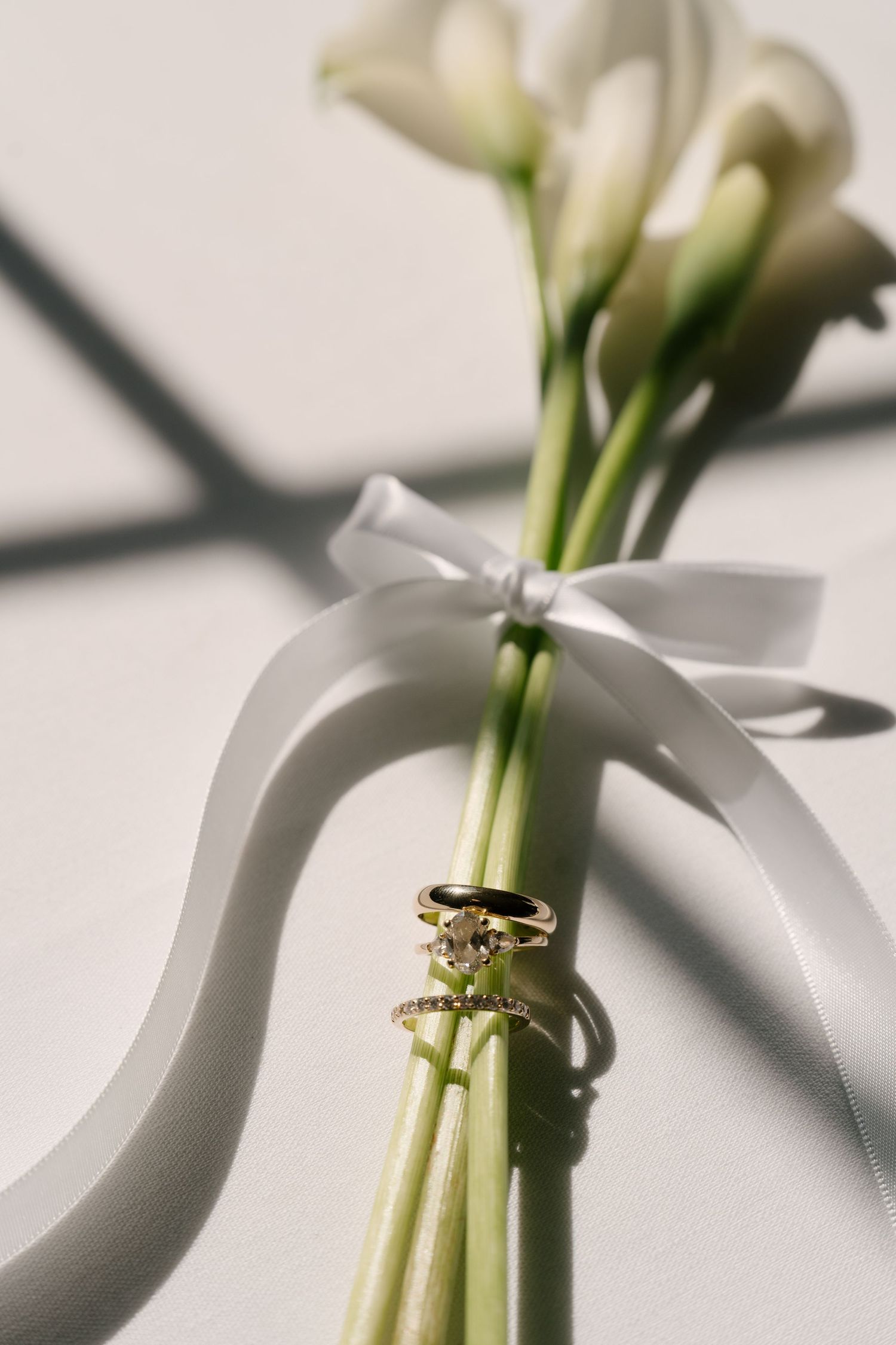 Gold engagement and wedding rings resting on white calla lily stems tied with a satin ribbon bow.