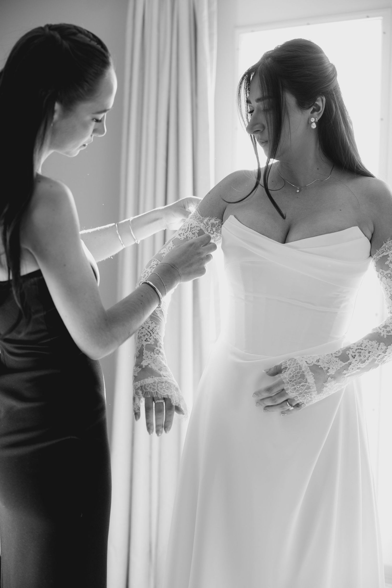 Bridesmaid in black dress adjusts lace sleeve of bride in white strapless gown, black and white.