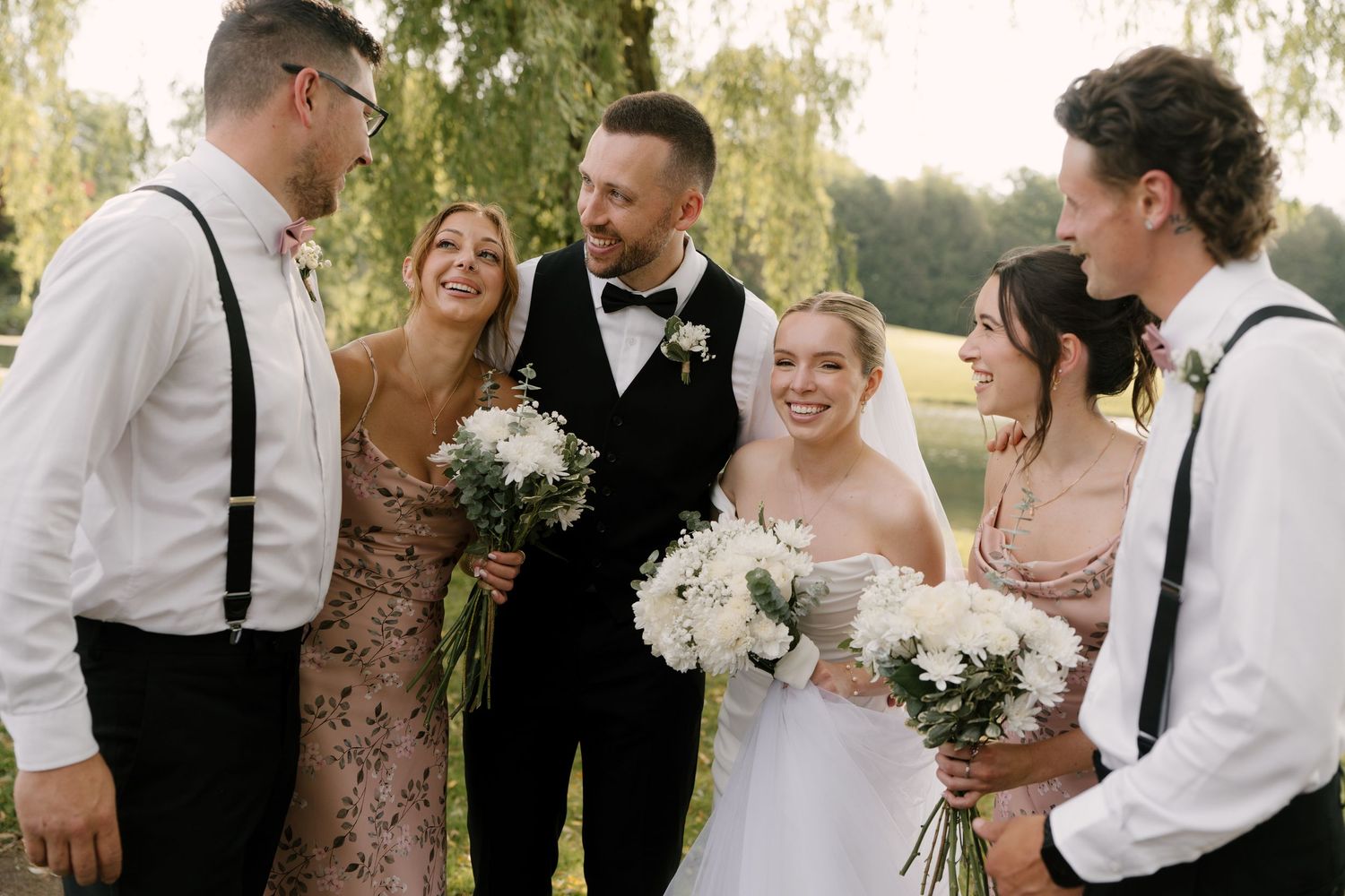 Bride and groom celebrate with wedding party guests outdoors, laughing and holding bouquets.