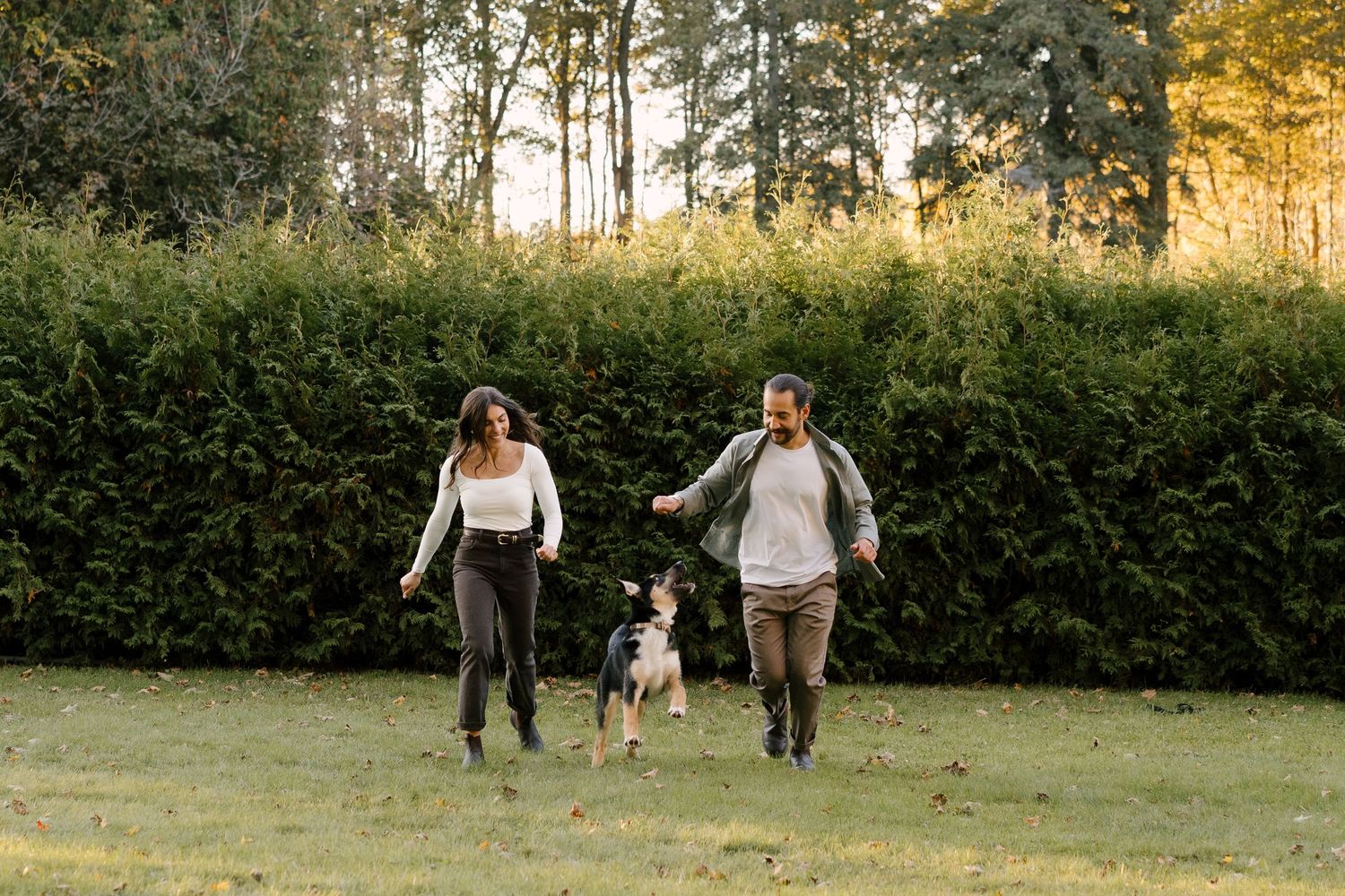 Couple running and laughing with their German Shepherd dog across a grassy park lawn.
