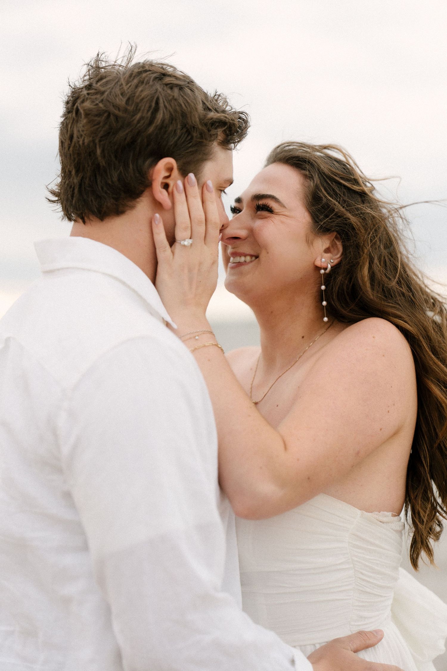 Bride with pearl earrings and engagement ring tenderly cupping groom's face on the beach.