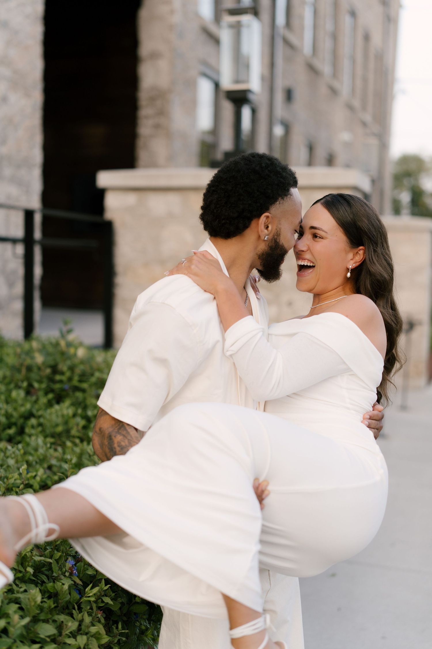 Man in white suit lifts laughing woman in off-shoulder dress outside a brick building.