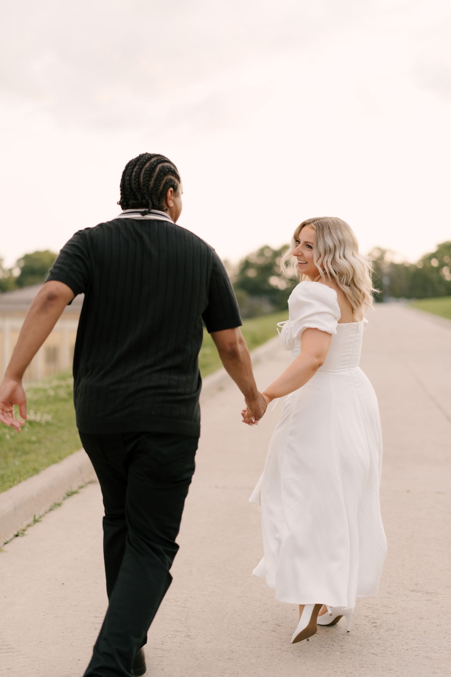 Couple walking hand in hand on a path, woman in white dress glancing back smiling at camera.