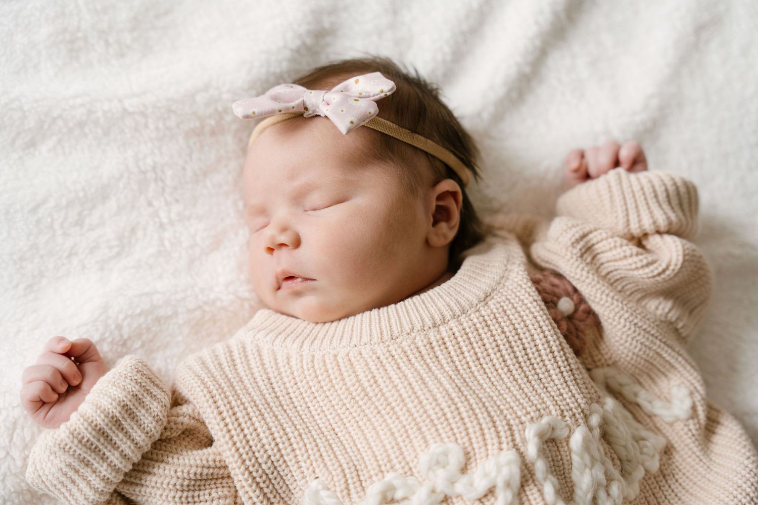 Peaceful newborn baby sleeping in cream chunky knit sweater with pink bow headband on white blanket.