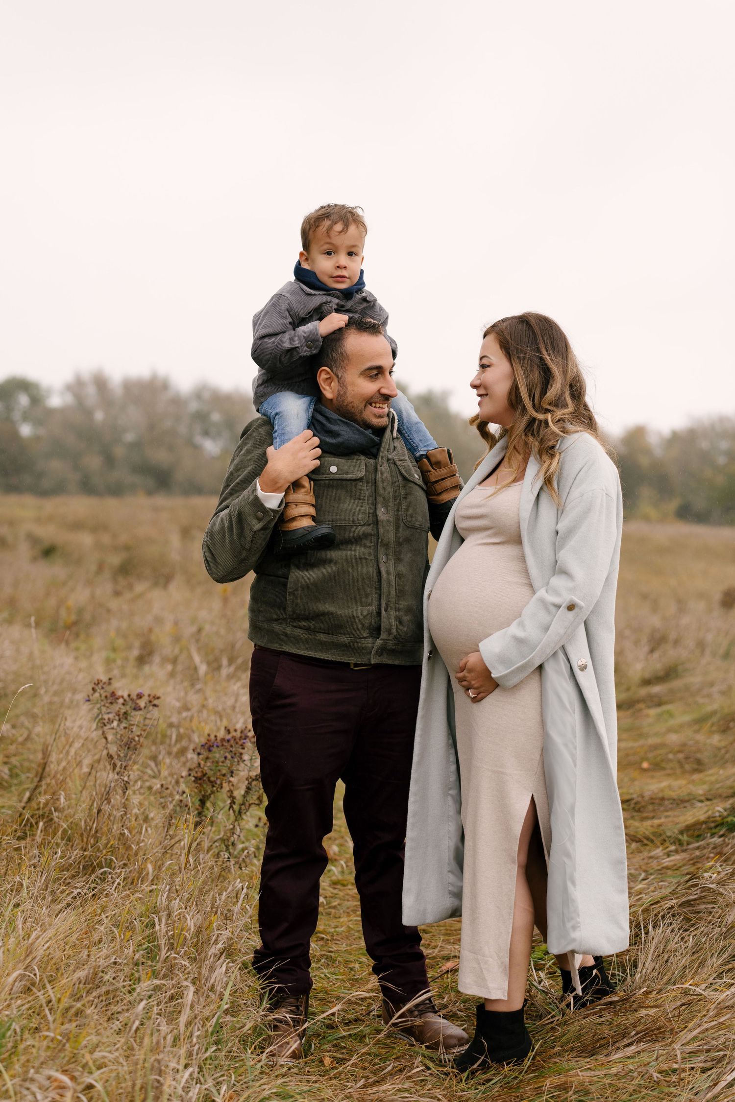 Smiling pregnant woman in grey coat with husband carrying toddler on shoulders in autumn field.