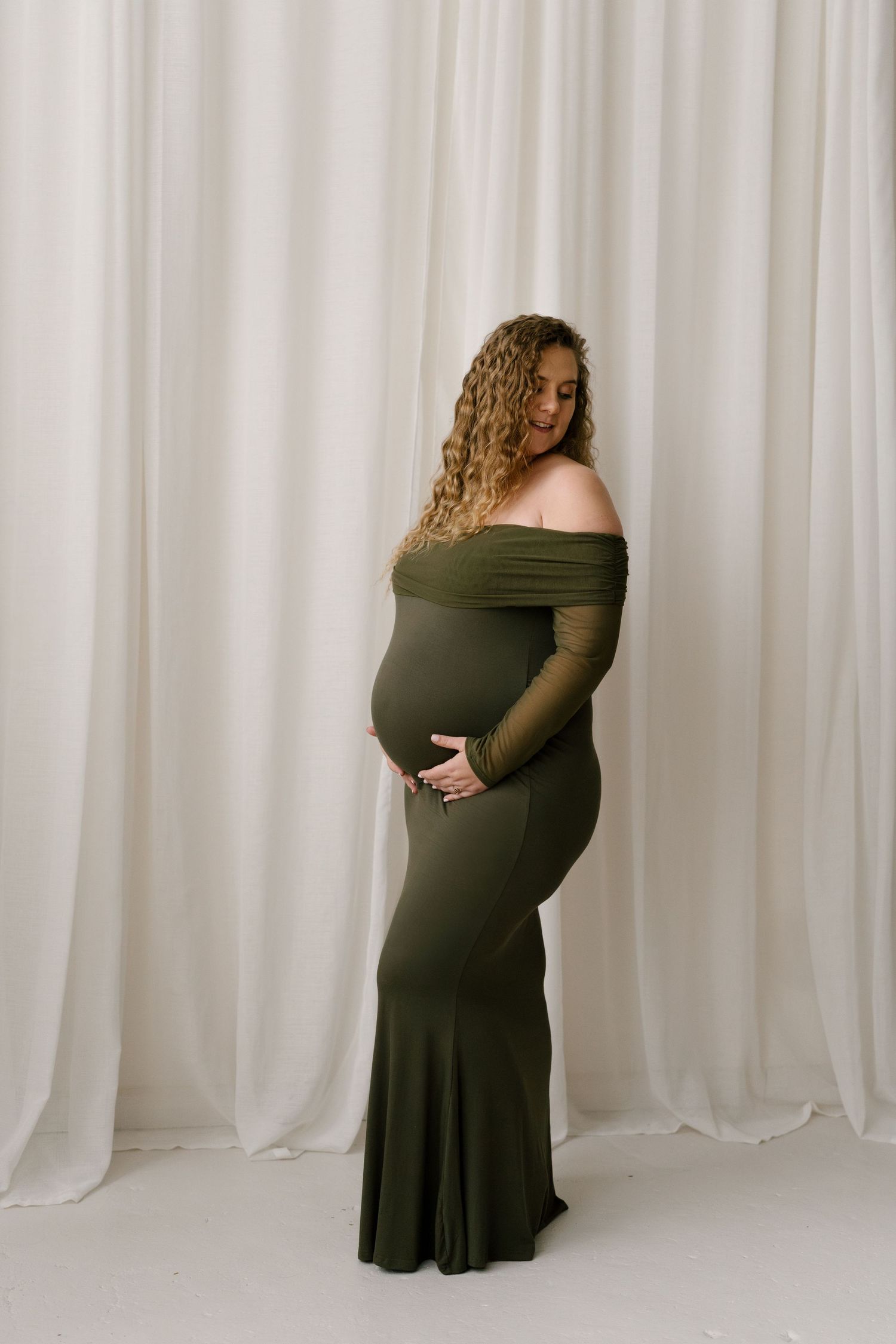 Pregnant woman in olive off-shoulder gown cradling bump against white curtain backdrop.