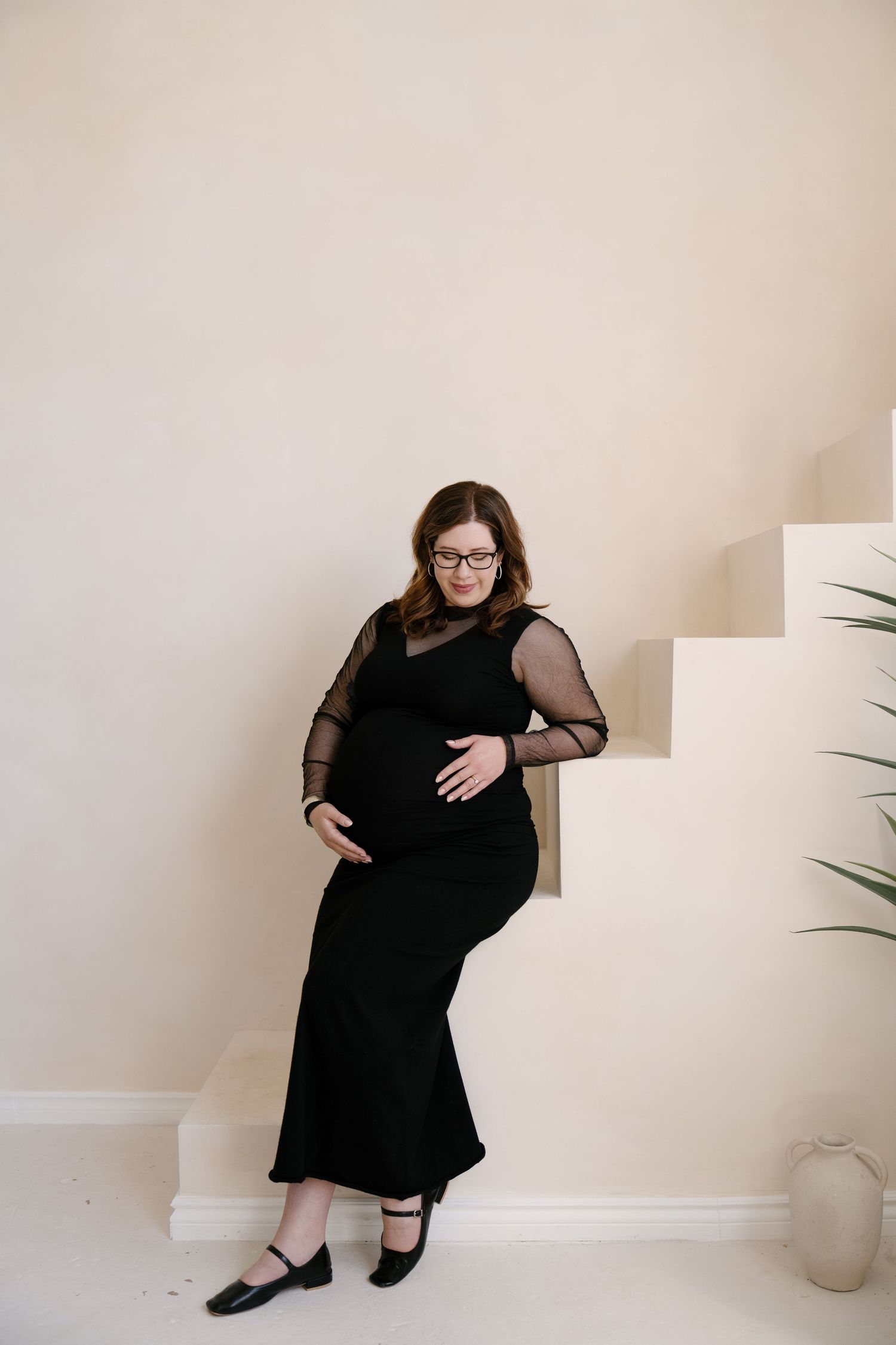 Pregnant woman in black mesh dress leaning against white staircase in a minimalist interior.