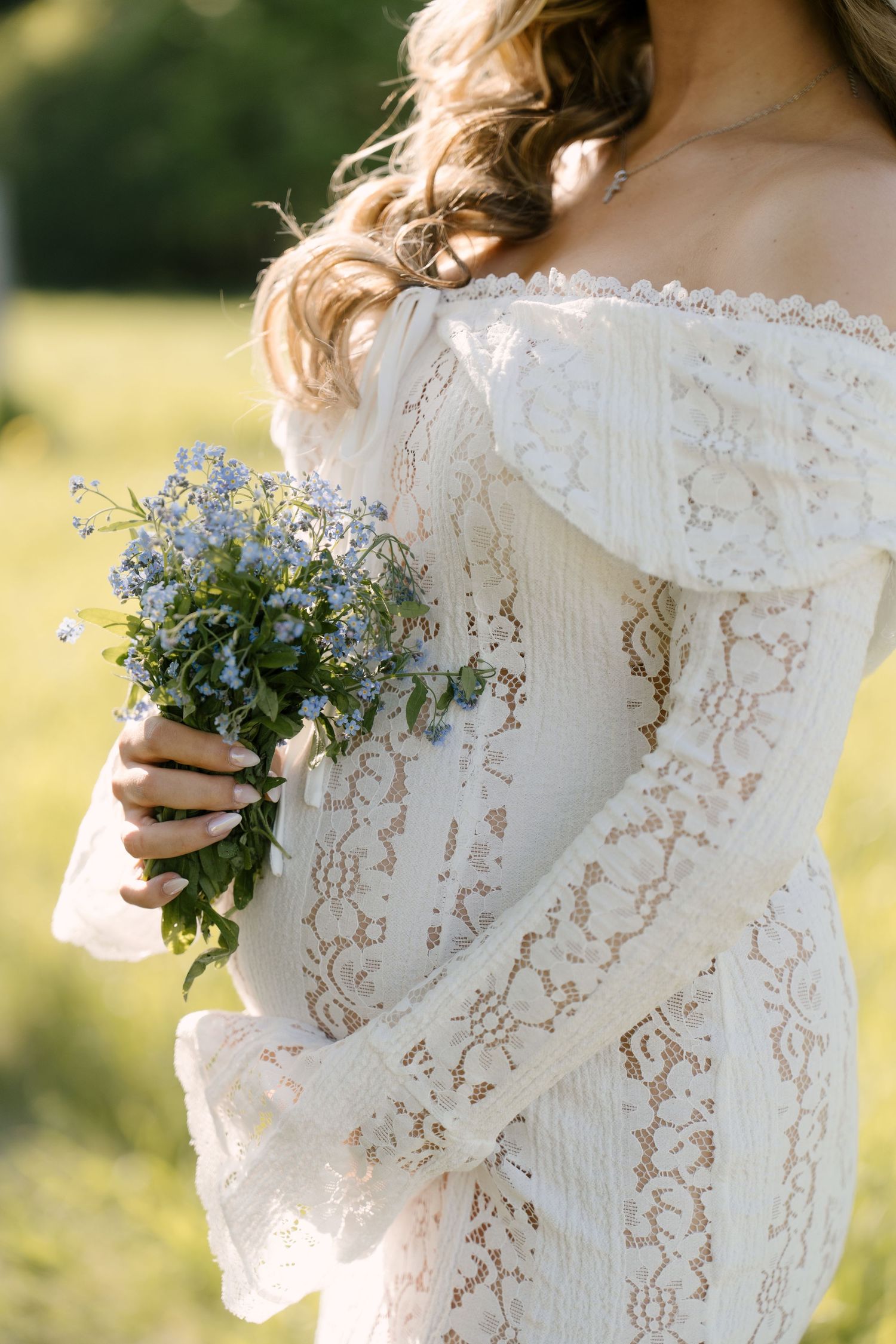 Pregnant woman in off-shoulder white lace dress holding a small bouquet of blue wildflowers outdoors.