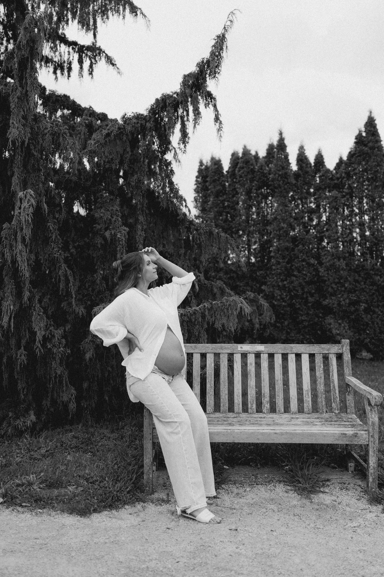 Black and white photo of a pregnant woman in white outfit leaning against a wooden bench outdoors.