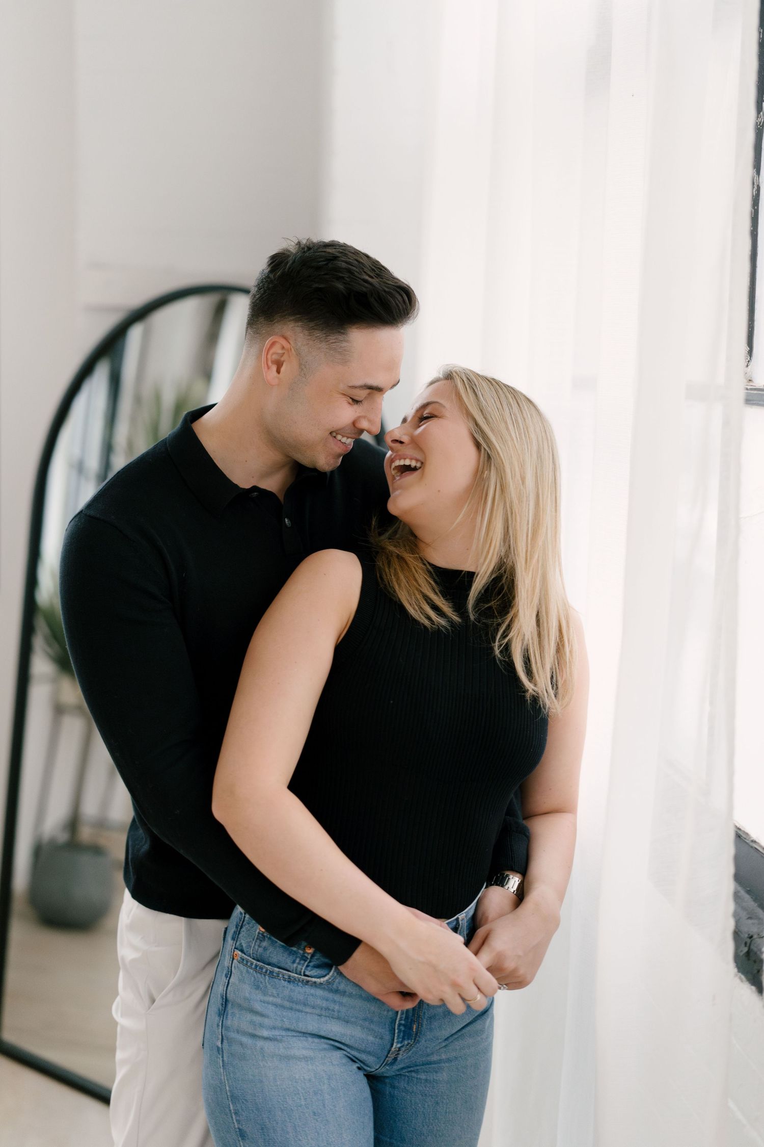 Laughing couple embracing closely near a large mirror in a bright white modern living space.