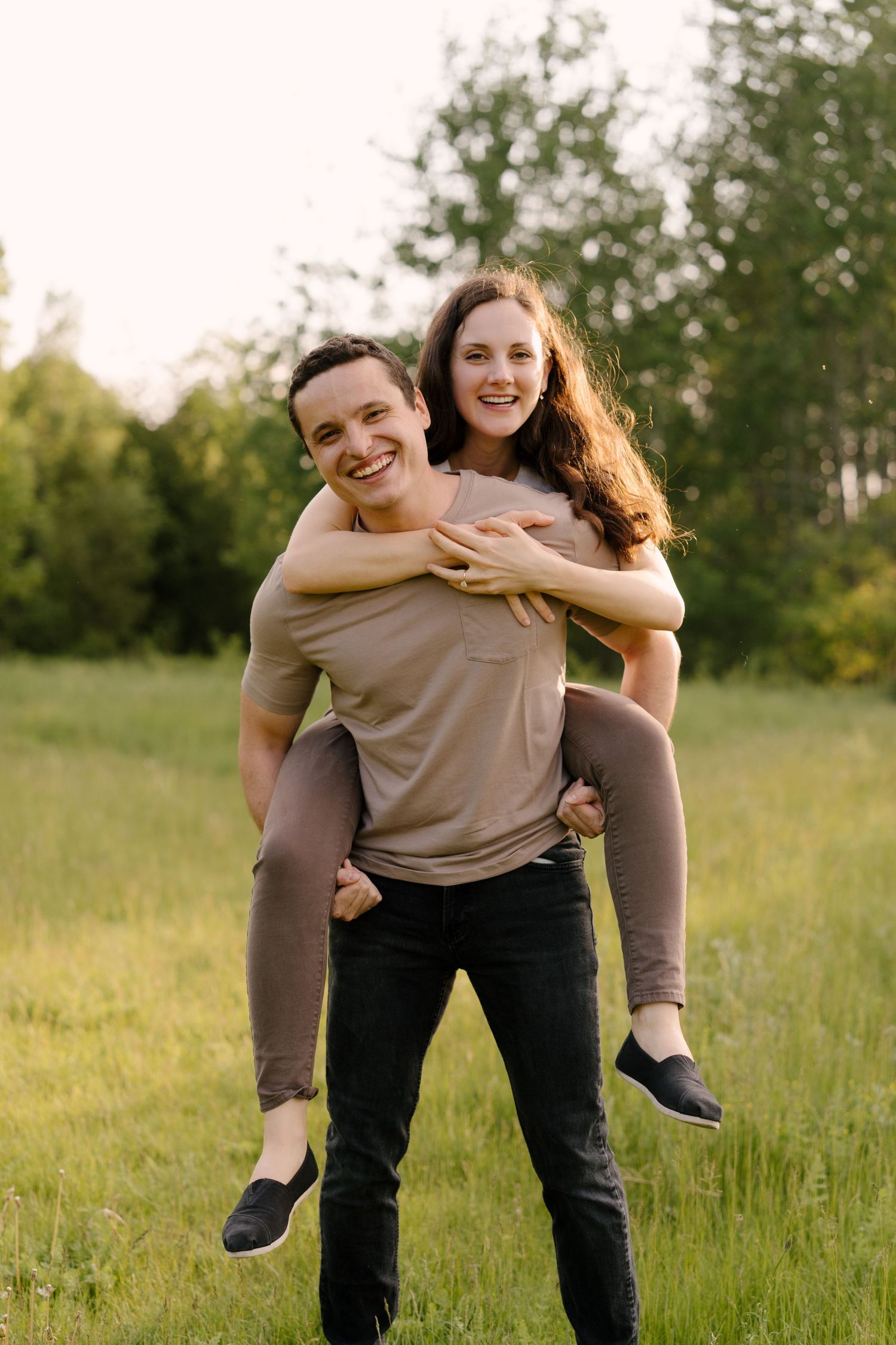Happy couple laughing in a grassy field at golden hour, man carrying woman on his back.