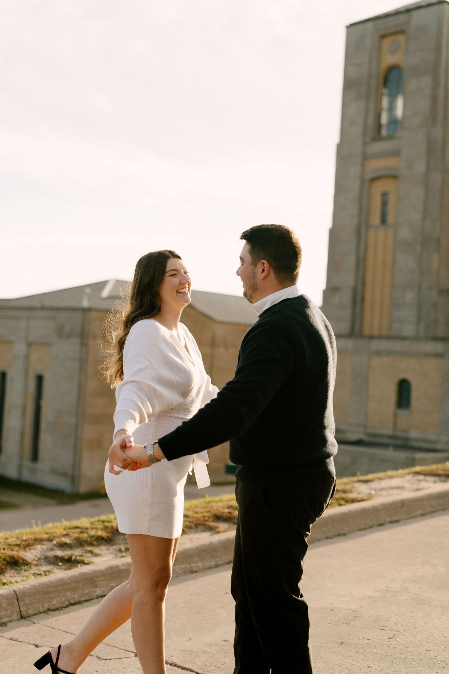 Smiling couple holding hands facing each other near a stone tower at sunset.