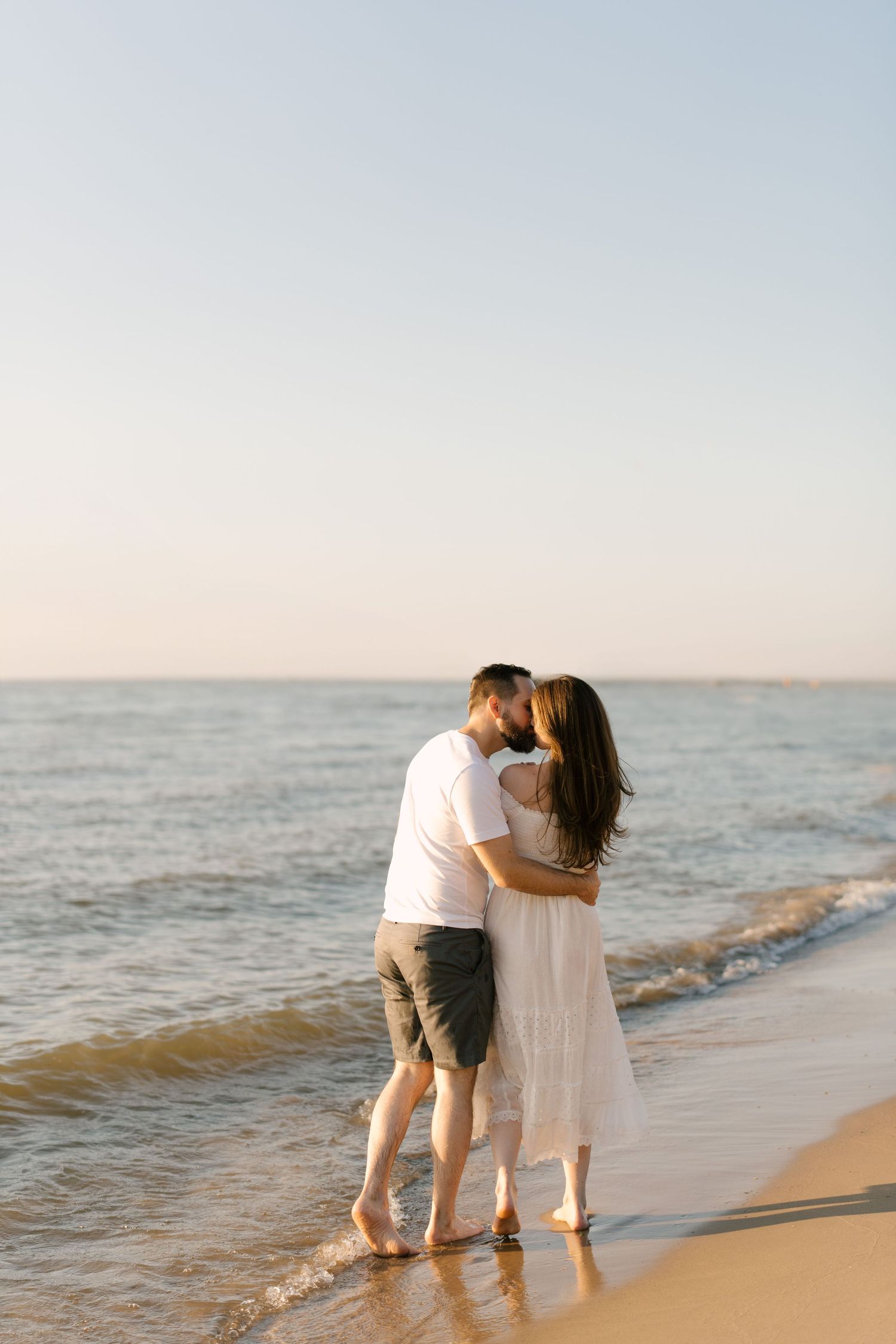 Romantic couple kissing on a sandy beach at sunset, embracing by the gentle ocean waves in soft golden light.
