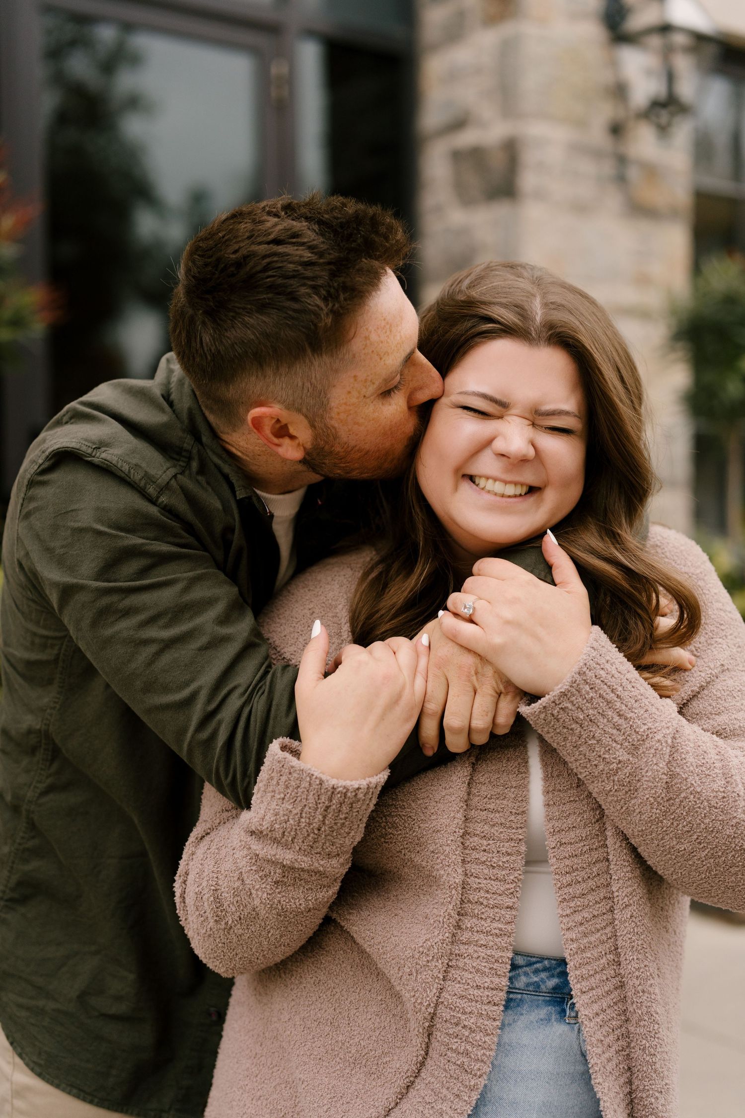 Man kissing woman on the cheek while embracing her from behind, both laughing outdoors.