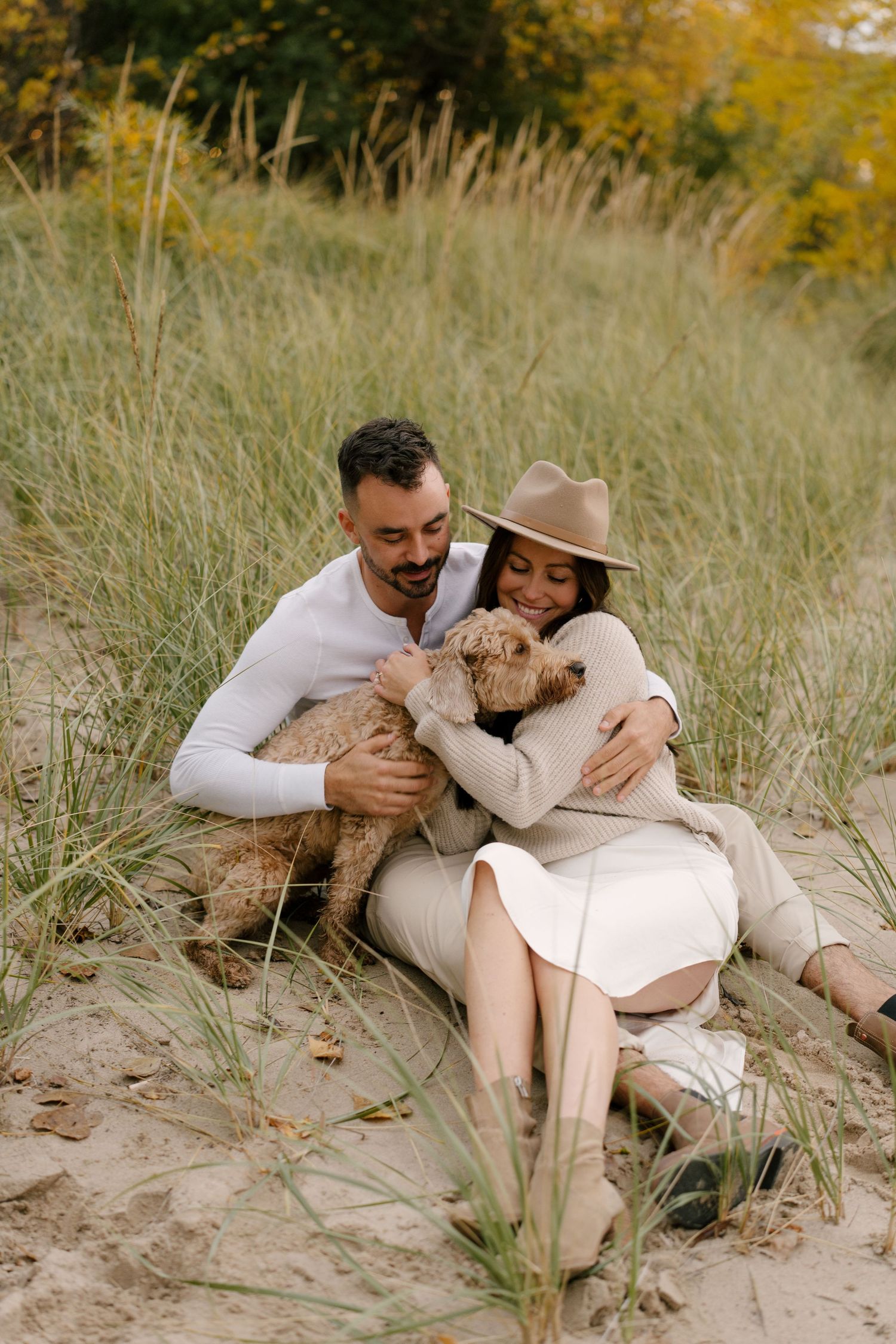 Couple cuddling goldendoodle dog while sitting in sandy beach grass dunes.