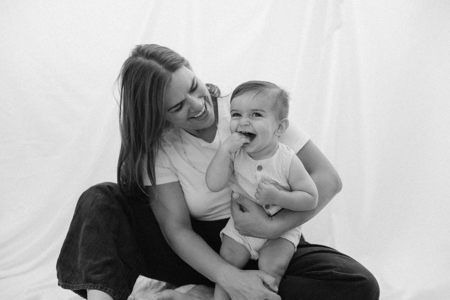 Black and white photo of a smiling young woman sitting with a happy laughing baby in a white onesie.