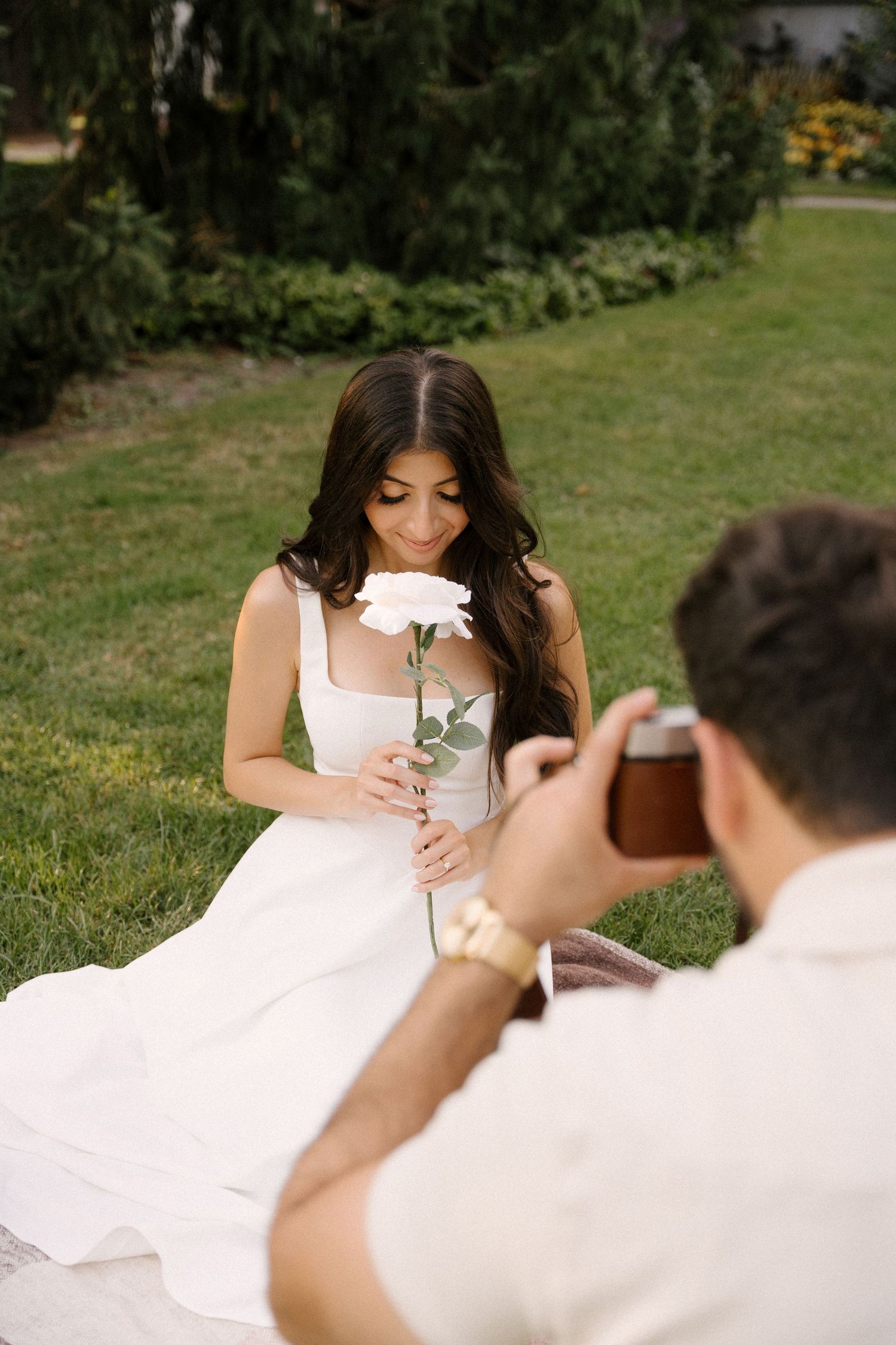 Woman in white dress smelling a white rose while partner photographs her on a garden blanket.