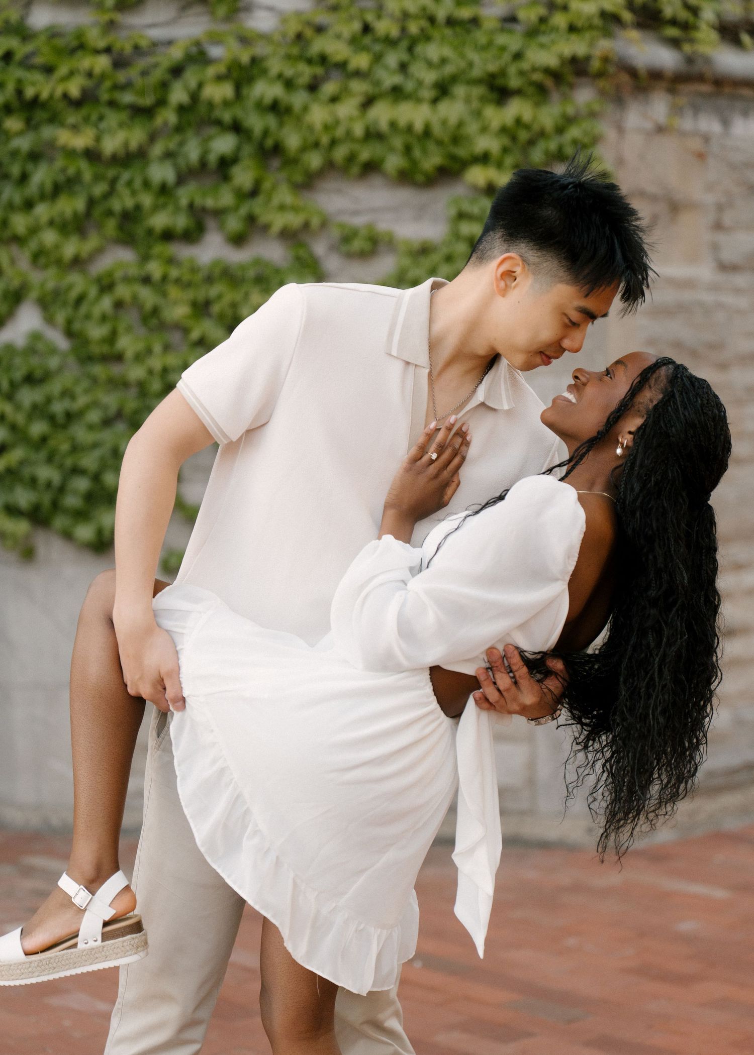 Man dipping woman in white dress while she smiles in front of an ivy-covered wall.