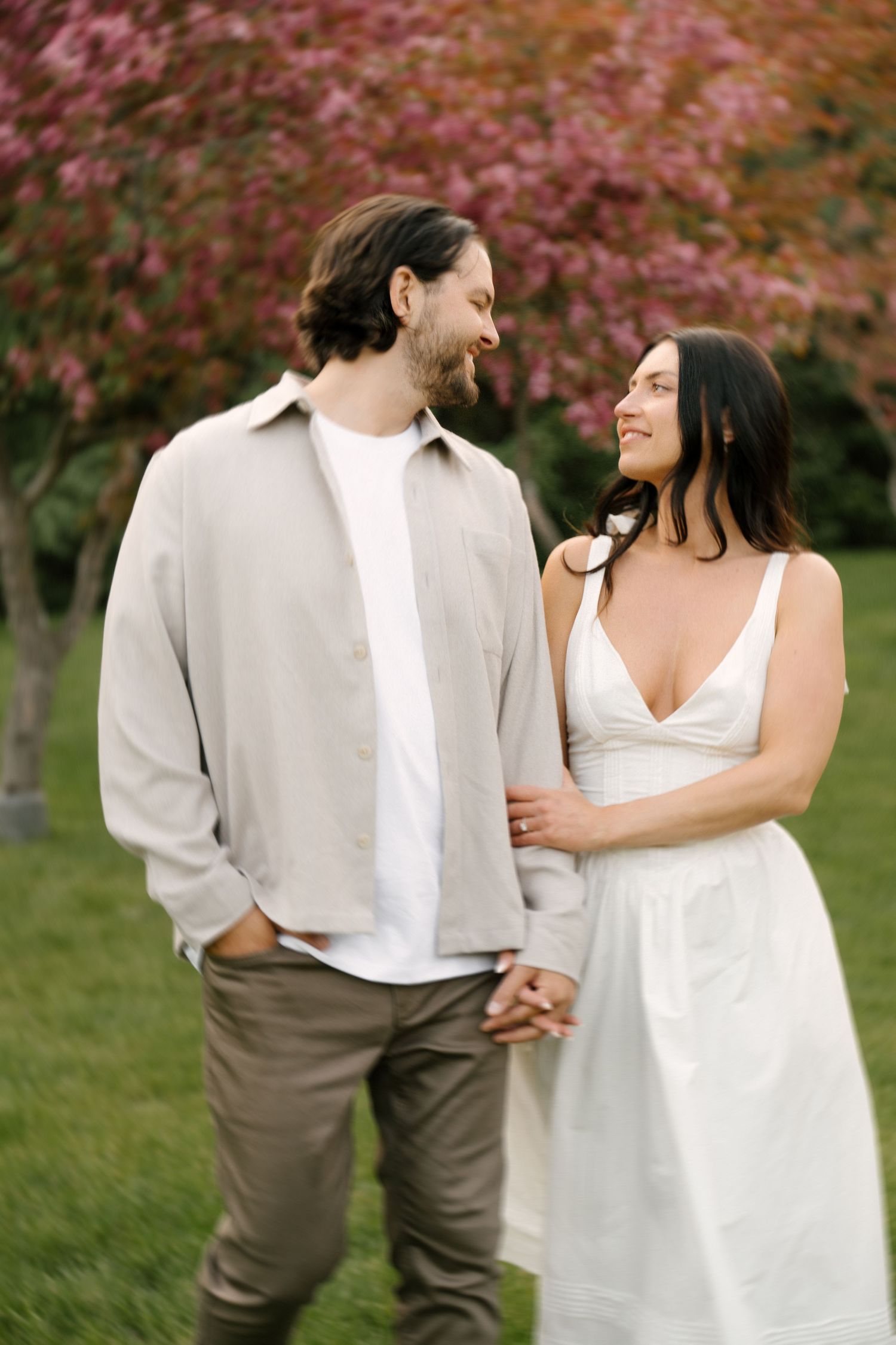 Smiling couple walking hand in hand through garden with pink flowering trees in background.