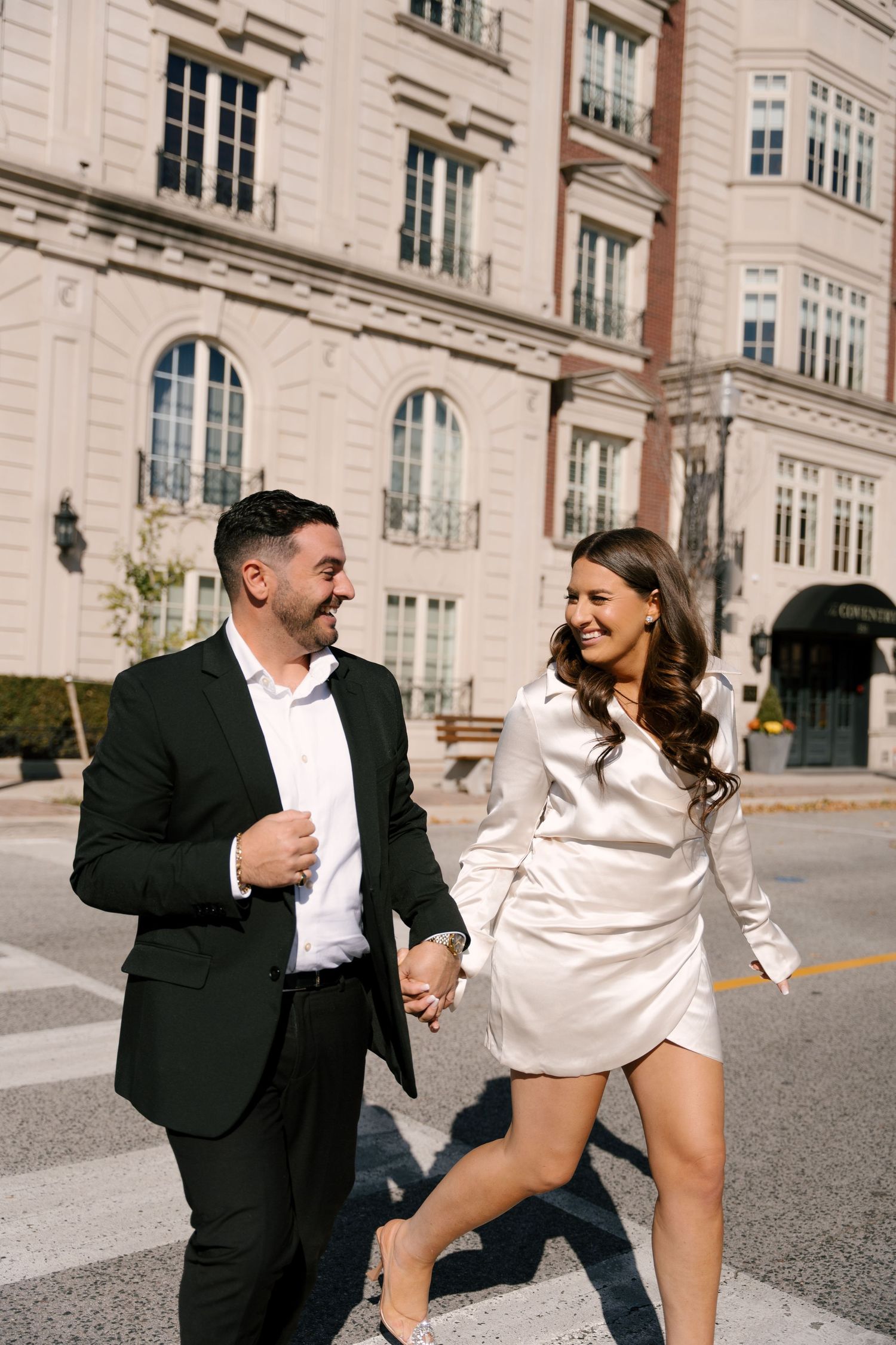 Laughing engaged couple crossing street hand in hand in front of grand urban building.