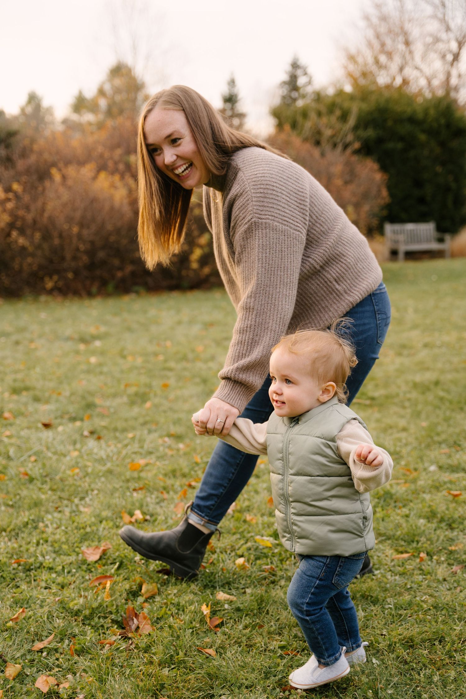 Cheerful mother helping smiling toddler in green vest walk on grass in autumn park.