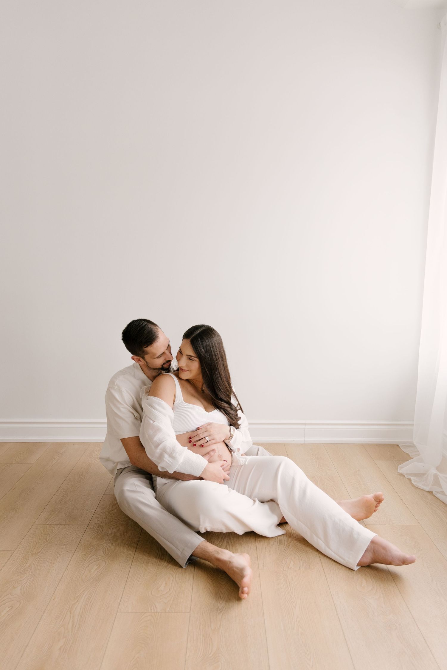 Couple in white outfits sitting on floor, man embracing pregnant woman from behind in bright minimalist room.
