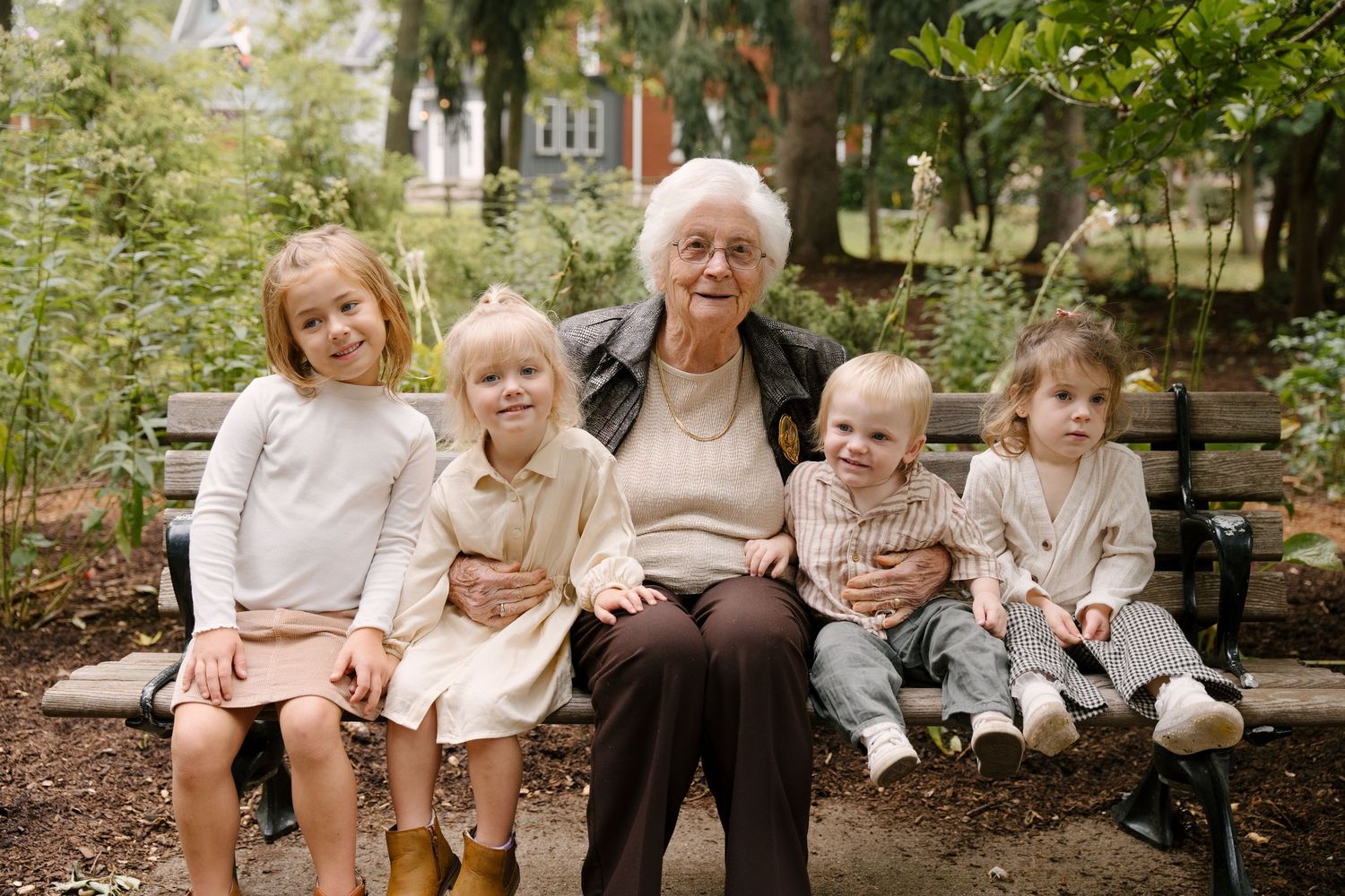 Elderly grandmother seated on bench surrounded by five young grandchildren in garden.