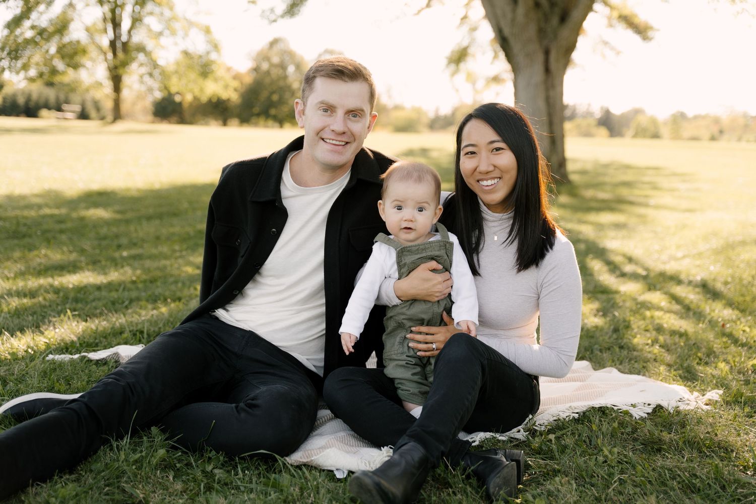 Happy interracial family with baby sitting on blanket outdoors under large tree.