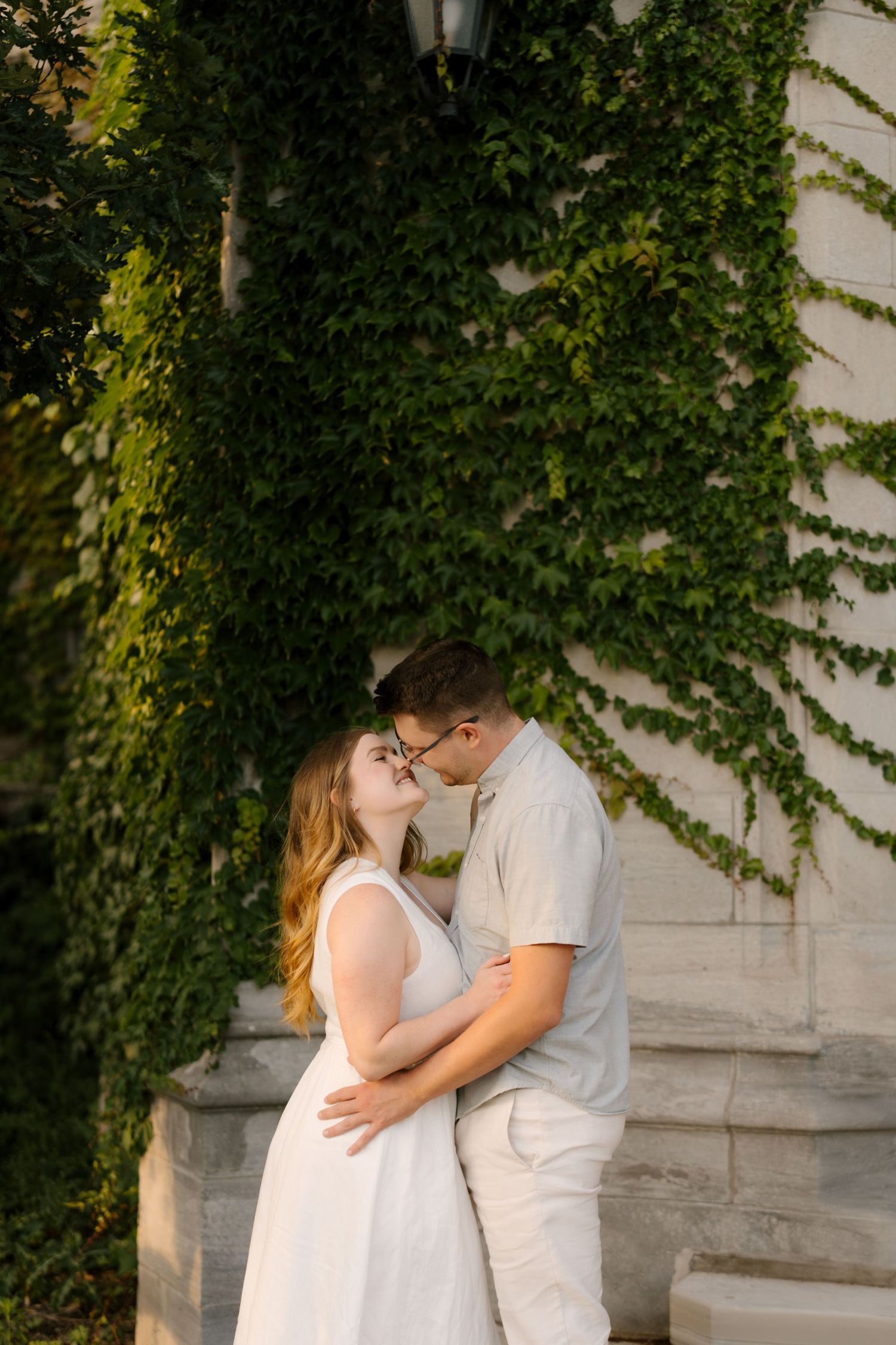 Couple sharing a kiss against ivy-covered stone building wall in warm evening light.